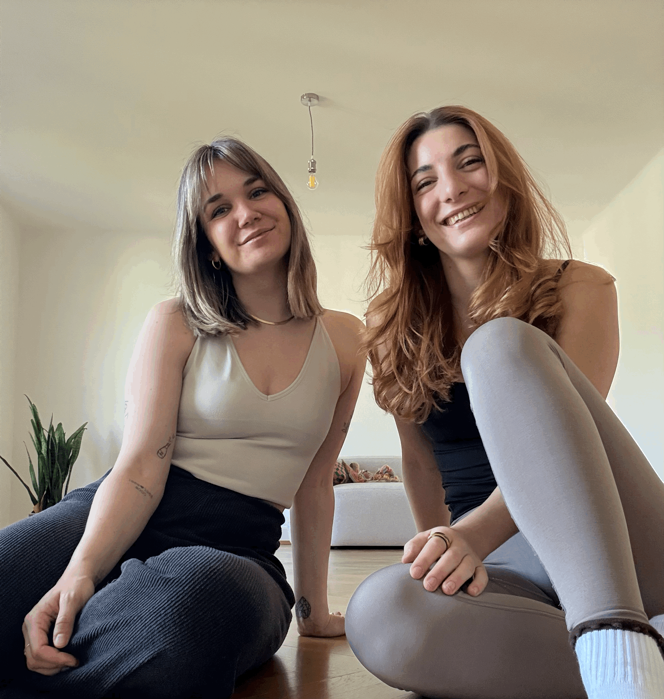Zwei junge Frauen sitzen auf dem Boden eines hellen Zimmers, lachen und posieren für die Kamera, während eine von ihnen auf einem großen runden grauen Teppich liegt.