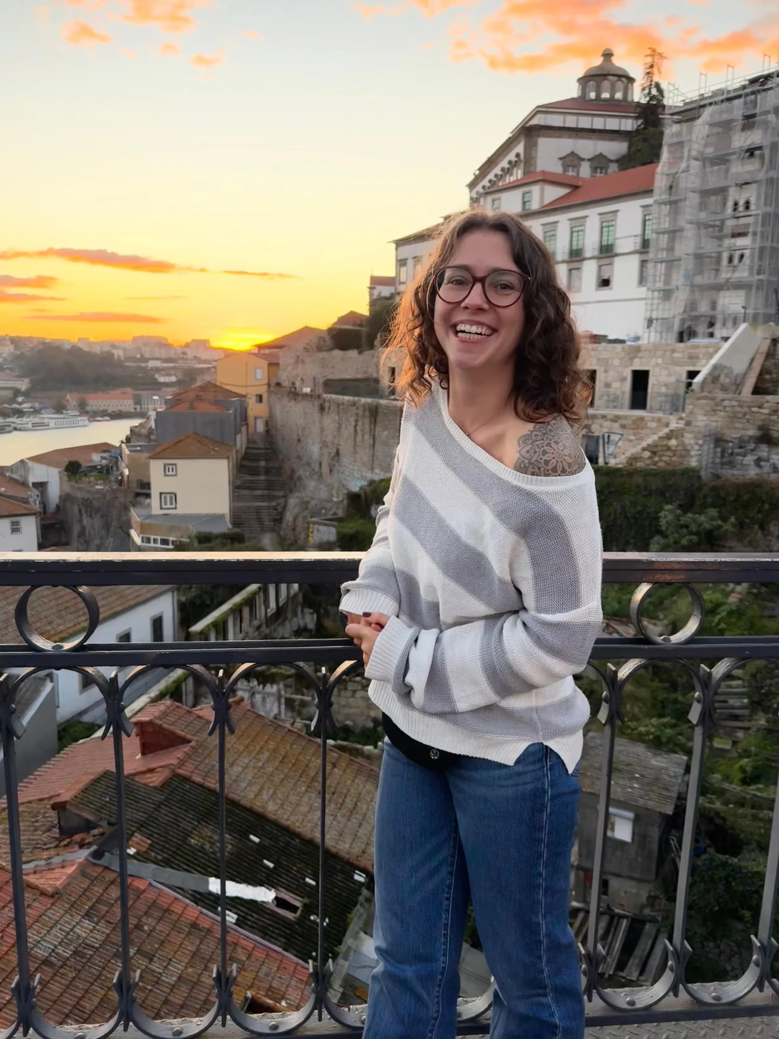 Junge Frau mit dunklen, lockigen Haaren und Brille, trägt ein weiß-gestreiftes Oberteil und blaue Jeans, steht auf einem Balkon mit Blick auf ein städtisches Panorama bei Sonnenuntergang, alte Gebäude mit roten Ziegeldächern im Vordergrund und ein Fluss im Hintergrund.