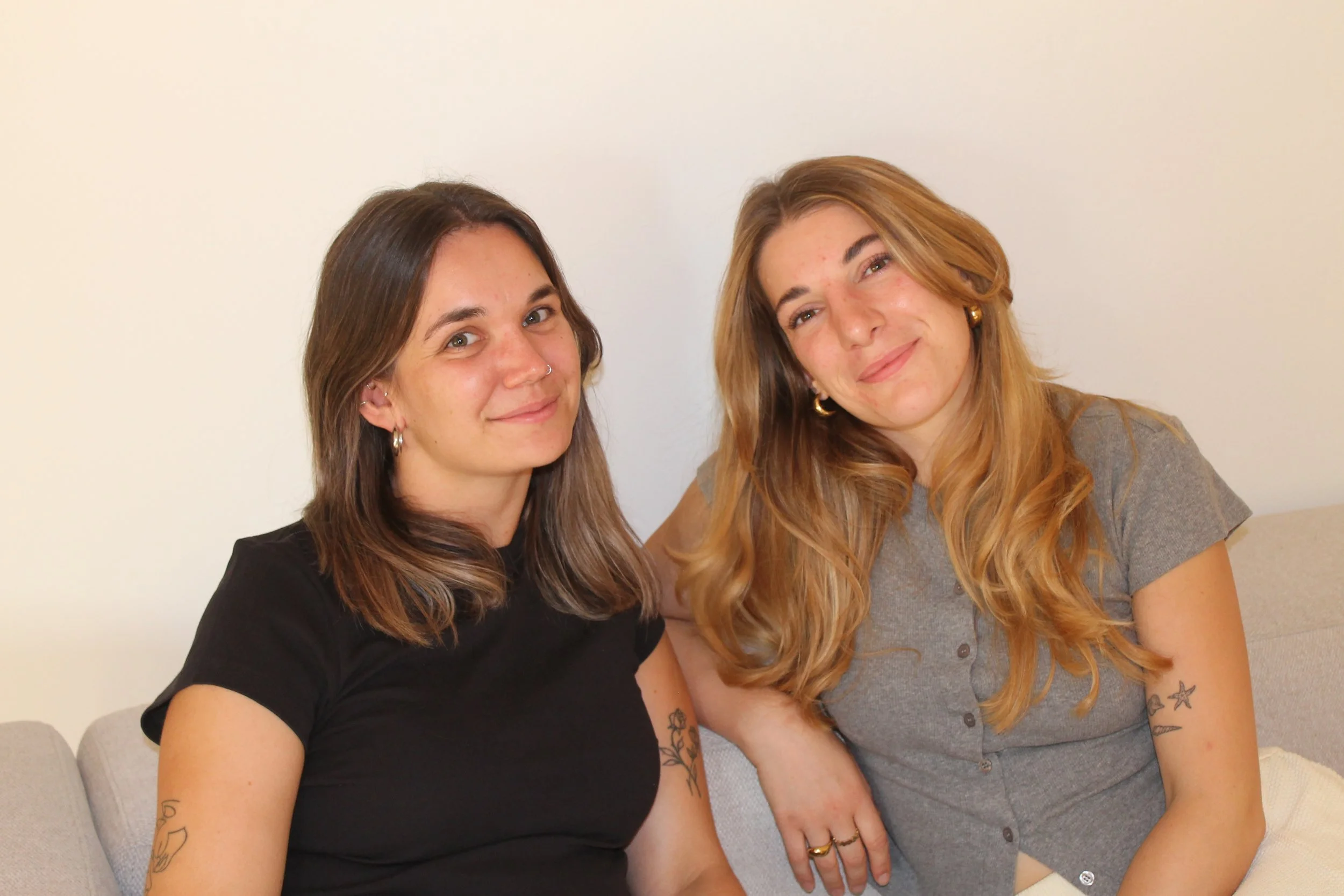 Zwei Frauen sitzen nebeneinander auf einer Couch vor einer weißen Wand, lächeln und schauen freundlich in die Kamera.