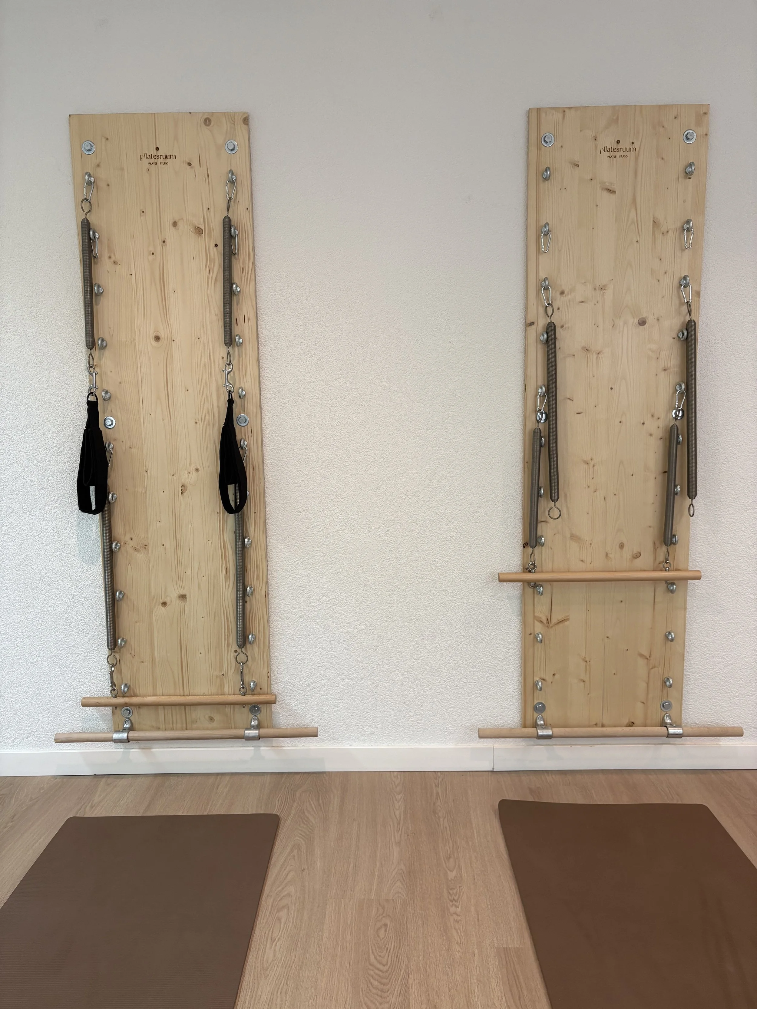 Zwei Wallboards bzw. Springboards mit Federn, Schlaufen und mit Pilates-Equipment, darunter Balance-Boards und Trapeze, befinden sich an einer weißen Wand. Vor den Paneelen liegen zwei beige Airex-Yoga-Matten auf einem Holzboden.