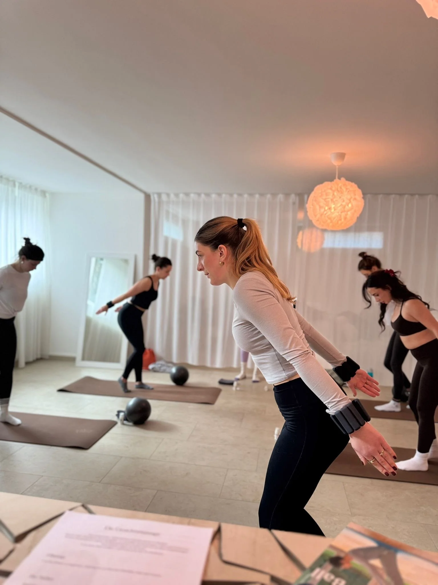 Frauen bei einem Pilates-Workout in einem hellen Raum mit Spiegel, Yoga-Matten und Medizinbällen.