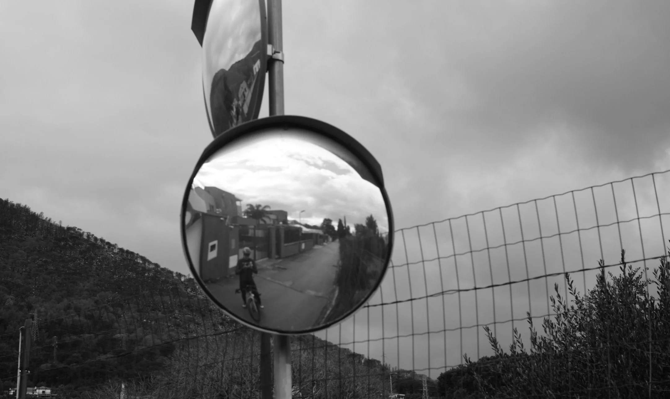 Black and white photo of a convex mirror on a street showing a reflection of a cyclist, with a fence and cloudy sky in the background.