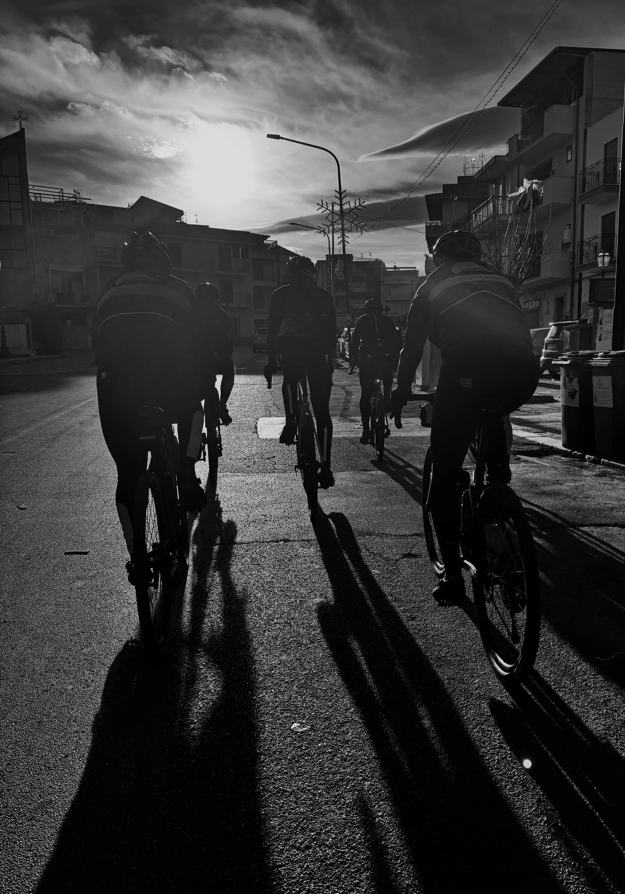 Black and white photo of cyclists riding on a street at sunrise, with long shadows on the road, and buildings in the background.