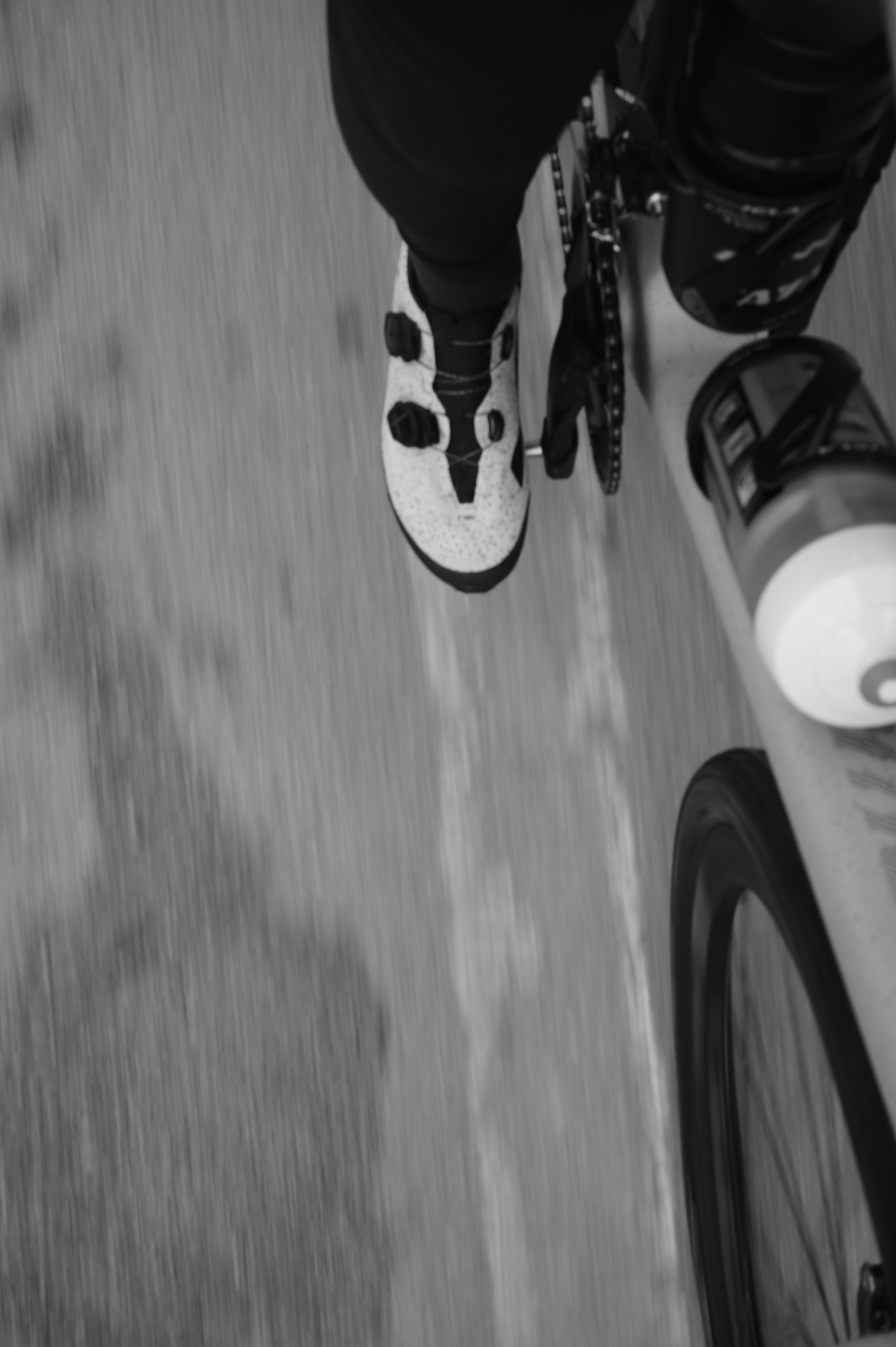 Cyclist's perspective of bike pedal and white cycling shoe, blurred motion, grayscale.