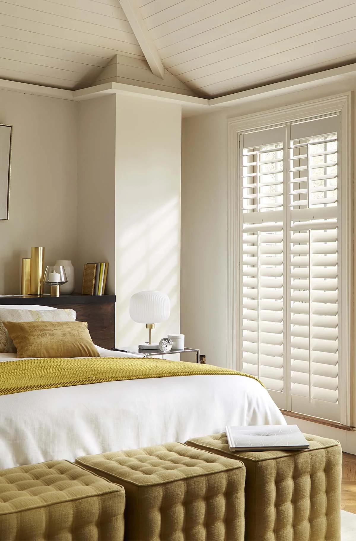 Bedroom interior with a bed, white linens, mustard-colored throw and pillows. Two mustard-colored ottomans at the foot of the bed. A side table with a white lamp and decorative items. White shuttered windows.