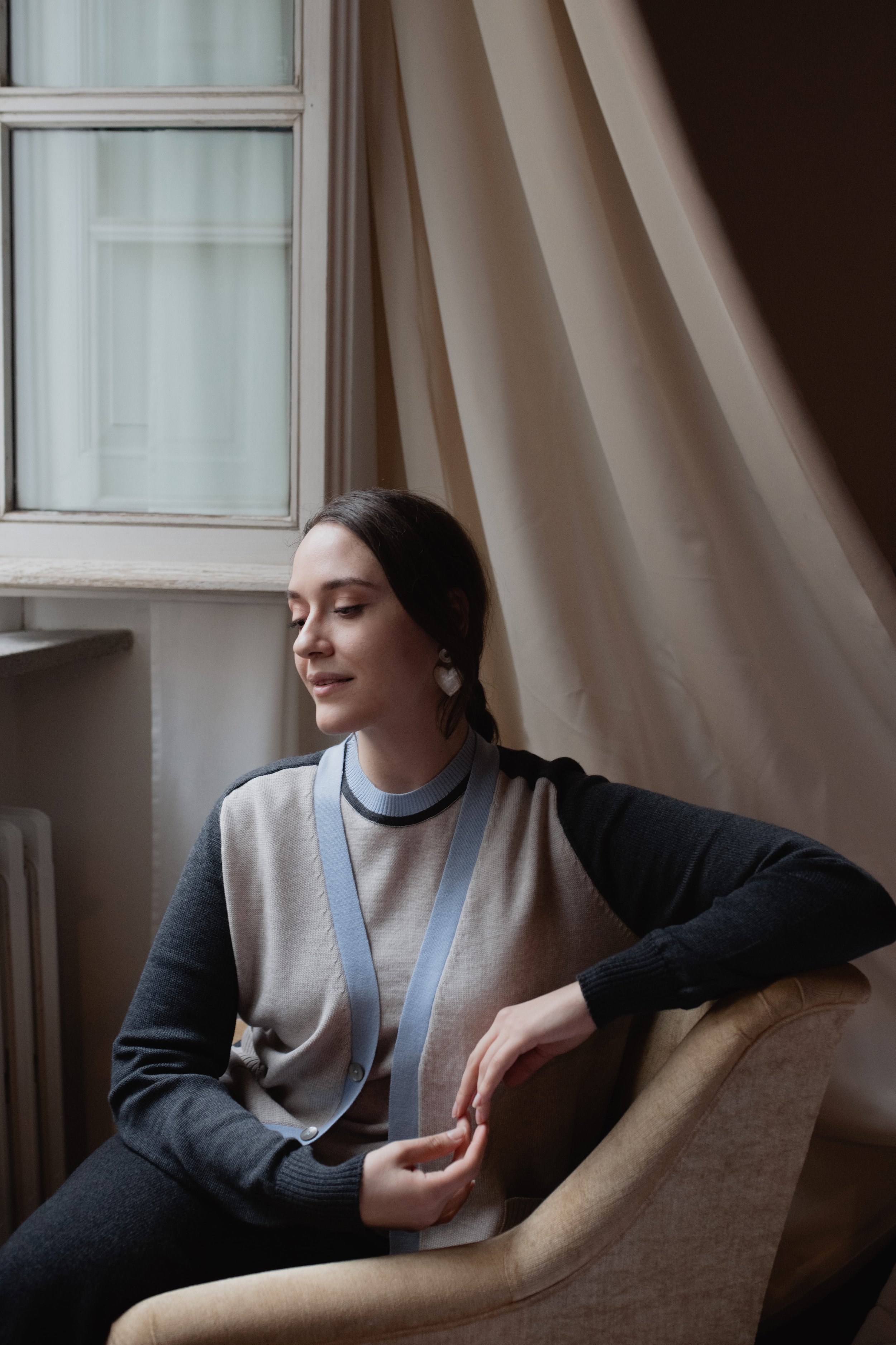 Donna seduta su una poltrona vicino a una finestra, indossa un maglione grigio e blu. Le tende bianche chiudono parzialmente la finestra.