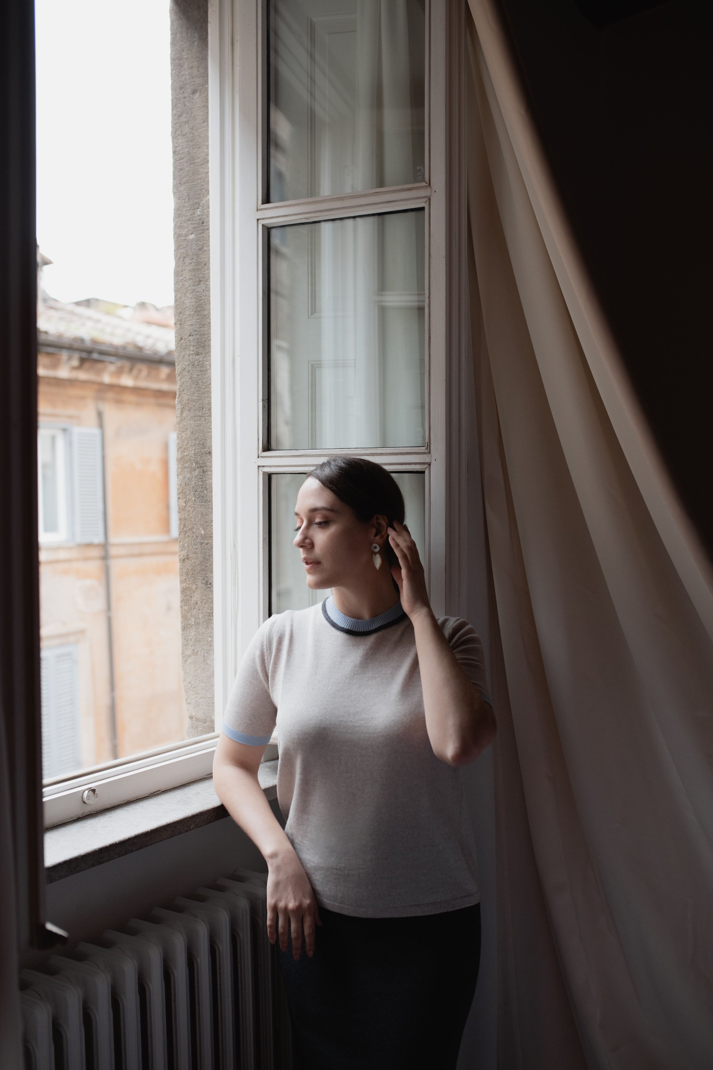Donna in piedi accanto a una finestra, con mano vicino all'orecchio, indossando una maglietta chiara. Sullo sfondo si vedono edifici esterni e una tenda interna.