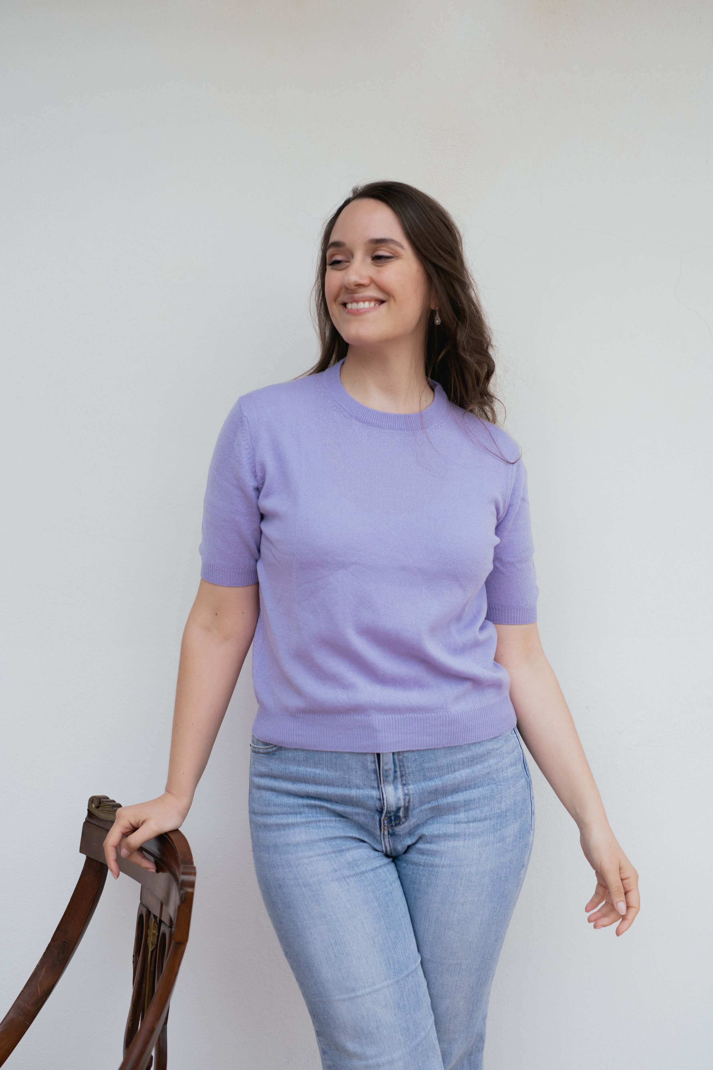 Donna sorridente con maglia viola e jeans chiari, appoggiata a una sedia di legno.