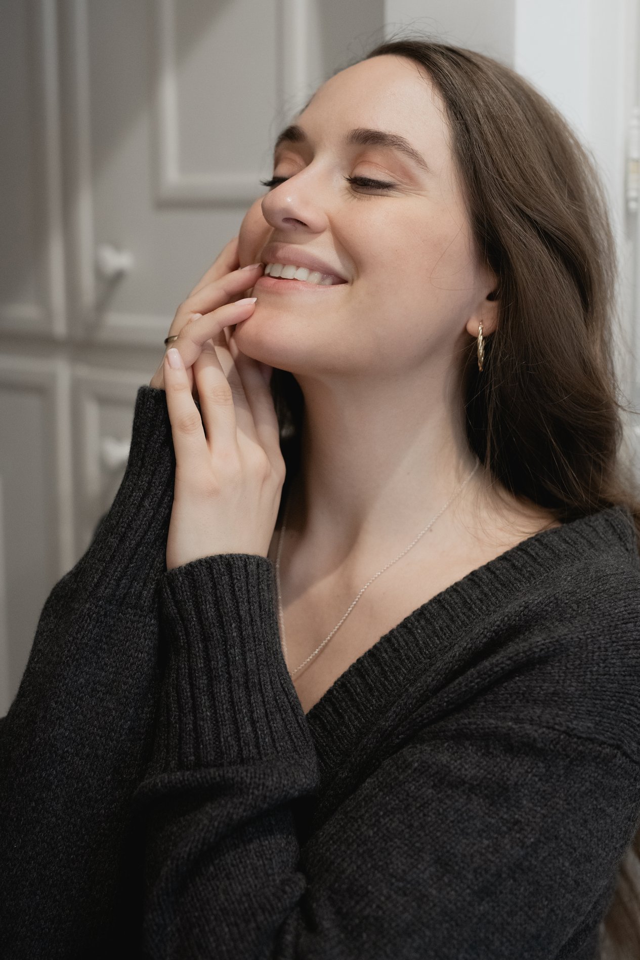 Donna sorridente con maglione scuro, occhi chiusi e mano sul viso in posa rilassata.