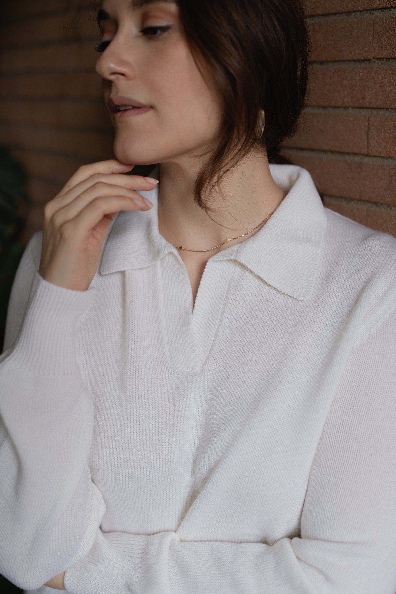 Donna con maglione bianco e collana dorata contro un muro di mattoni