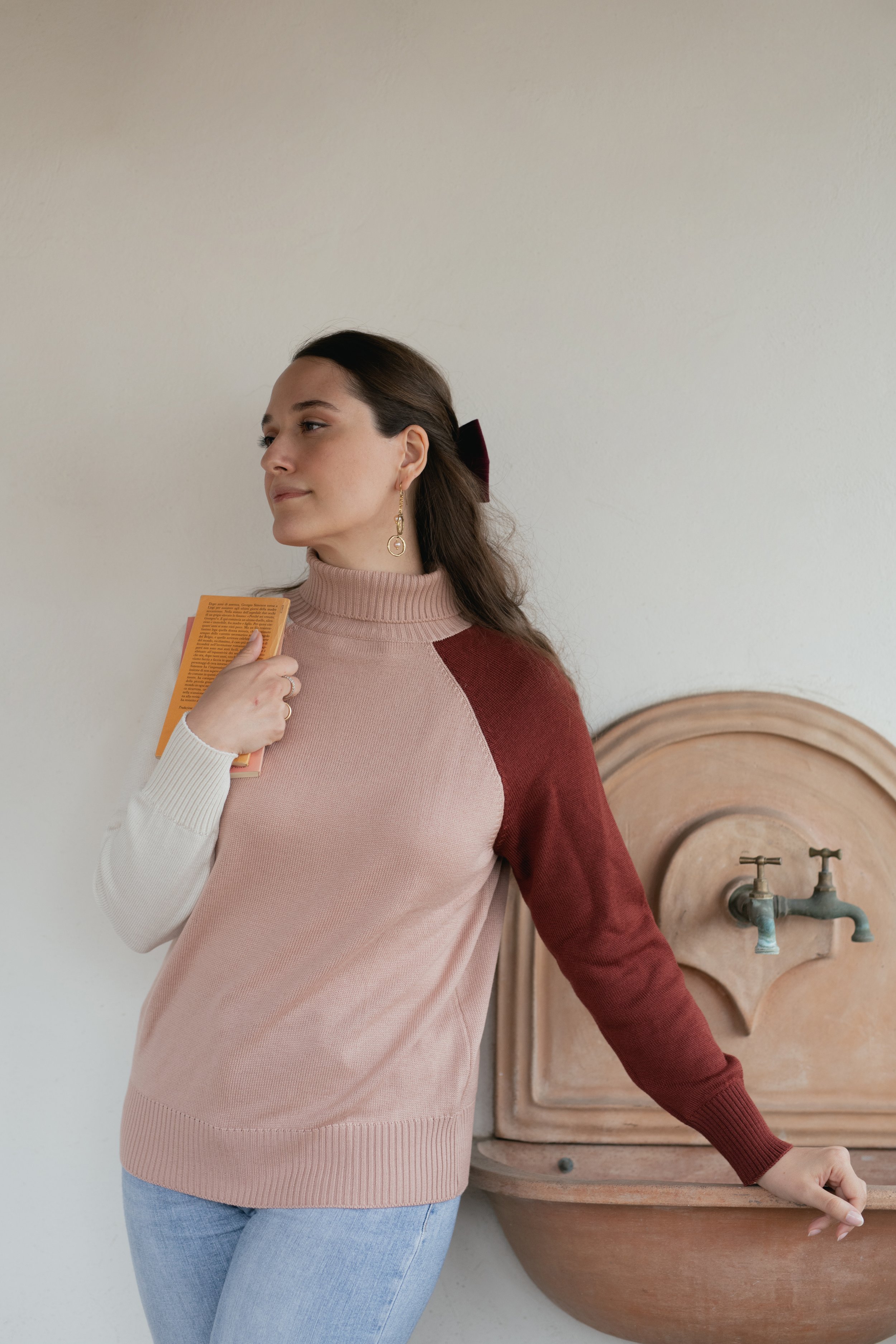 Donna con maglione bicolore regge un libro vicino a un rubinetto esterno.