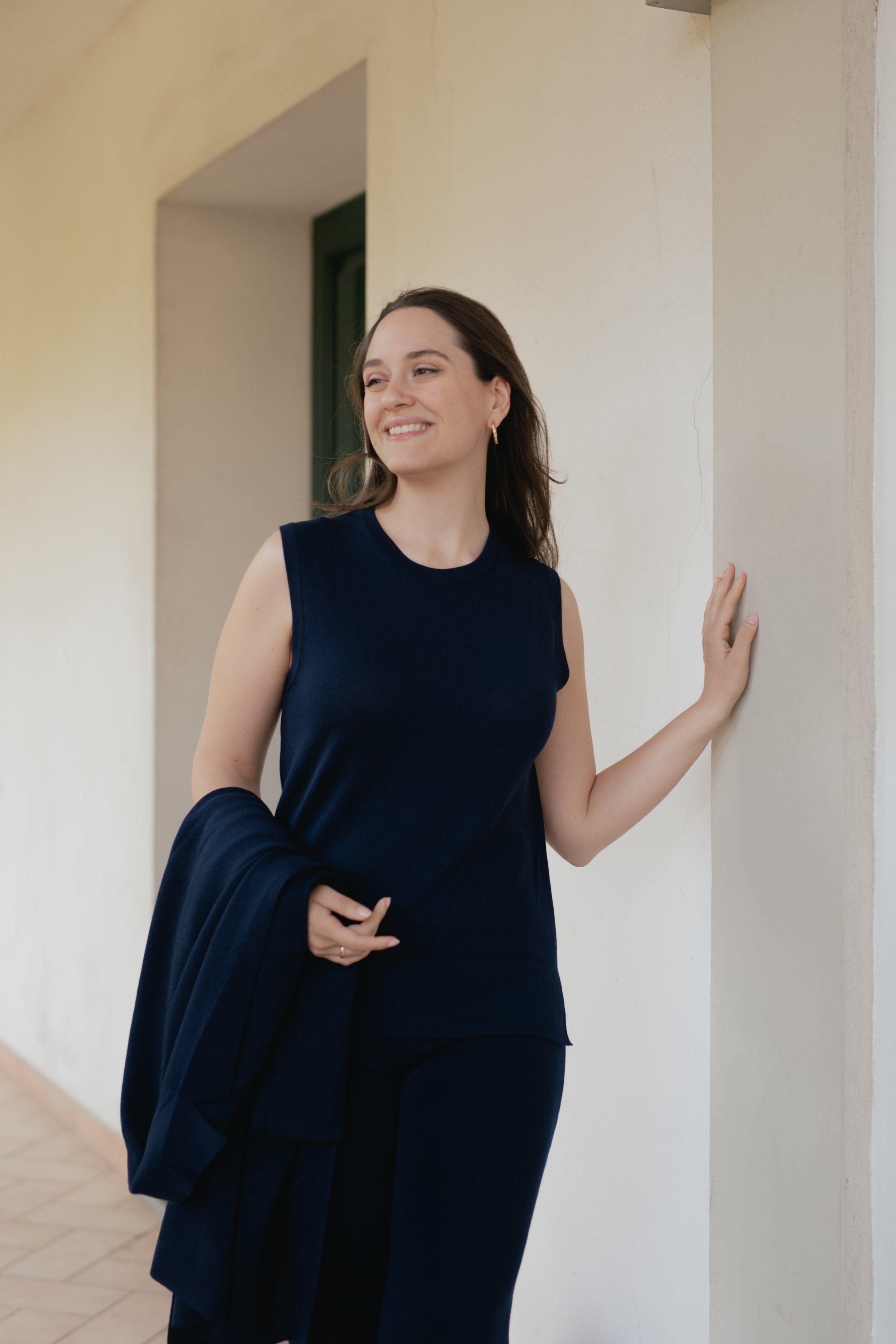 Donna sorridente in piedi vicino a un muro, indossando un abito nero senza maniche, con in mano una giacca. Sfondo di un corridoio con pavimento in piastrelle.