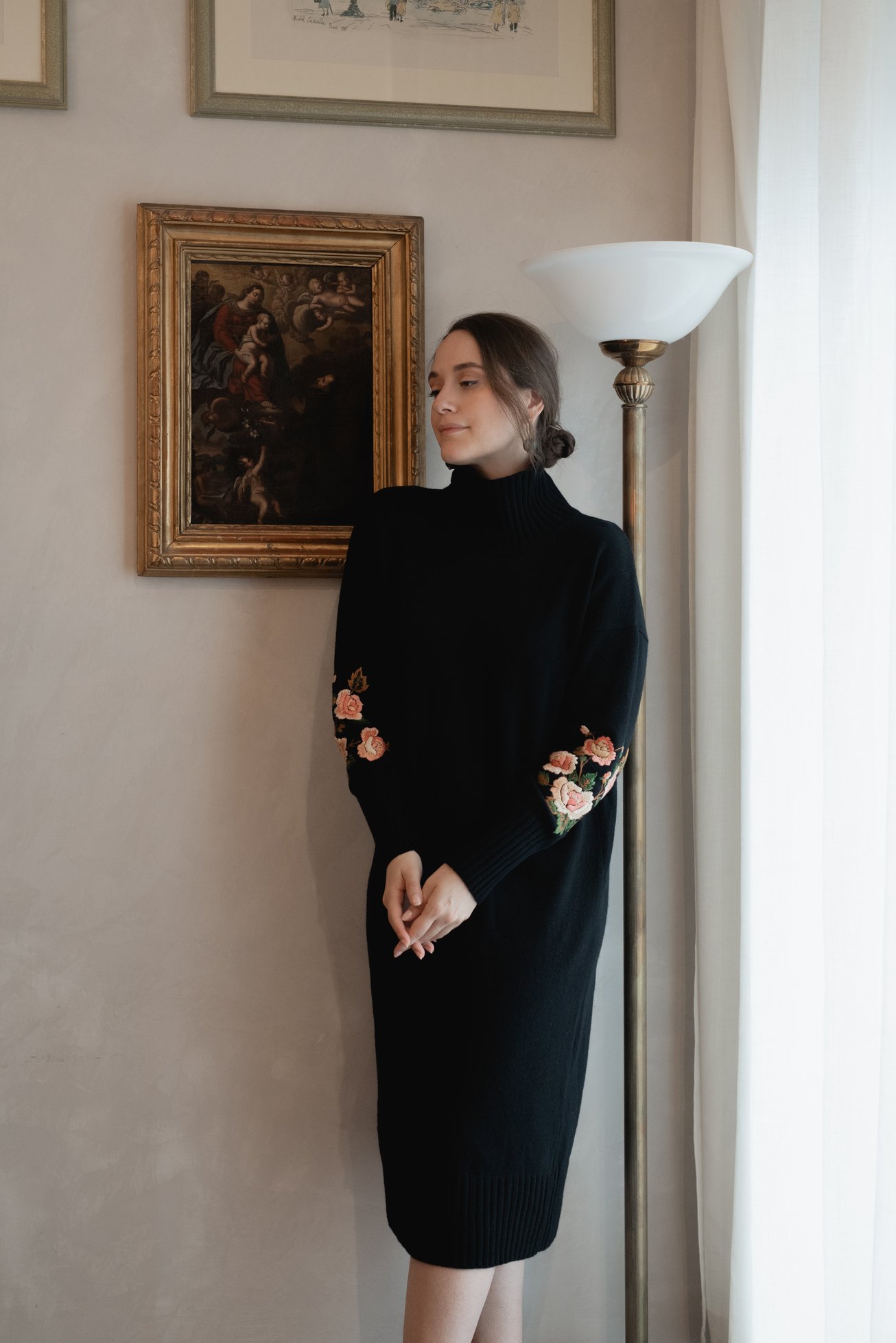 Donna in abito nero con fiori ricamati, in piedi accanto a lampada e quadro in cornice dorata su parete chiara.