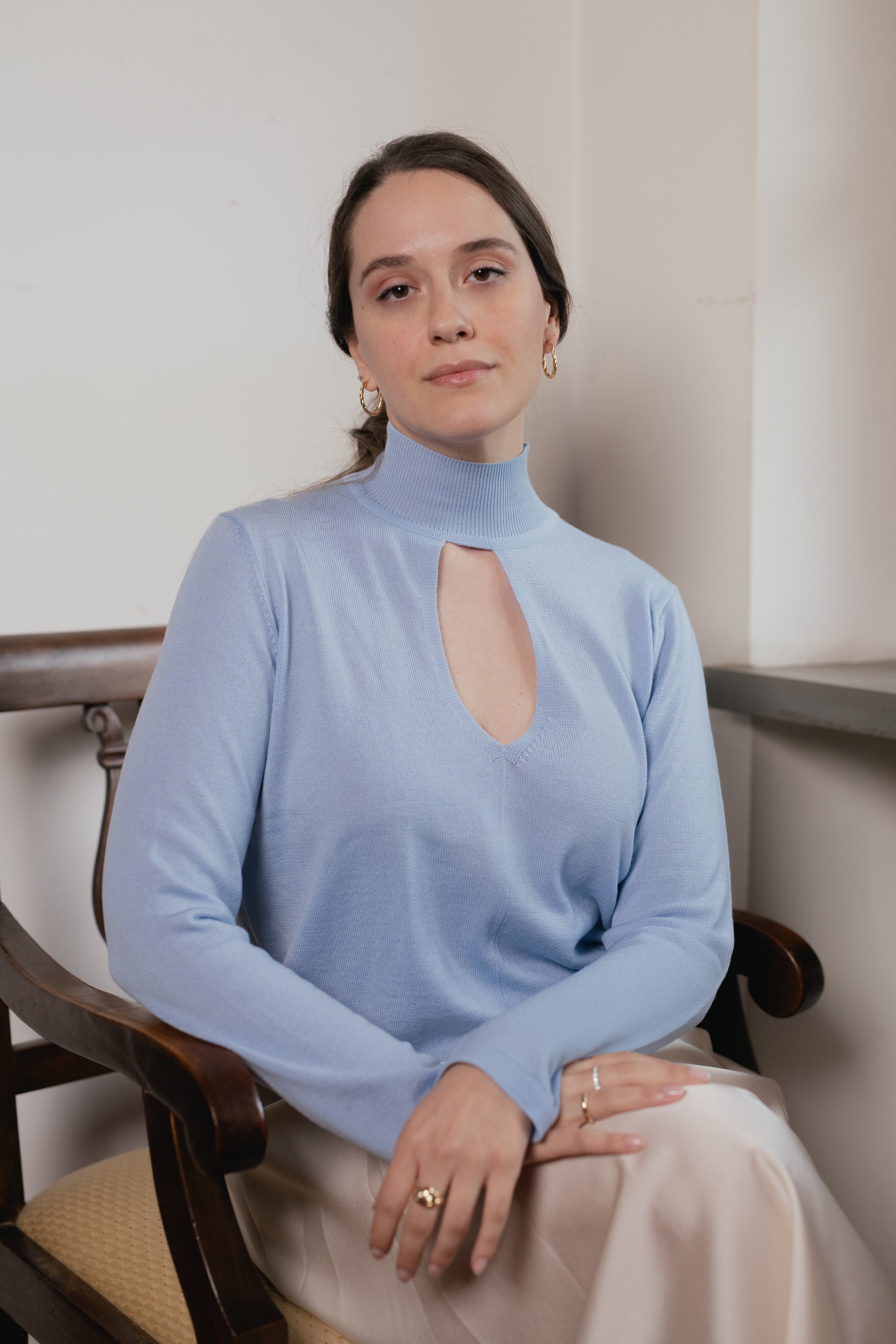 Donna seduta su una sedia indossando un maglione azzurro con scollo a goccia, capelli raccolti, orecchini a cerchio, e pantaloni chiari.