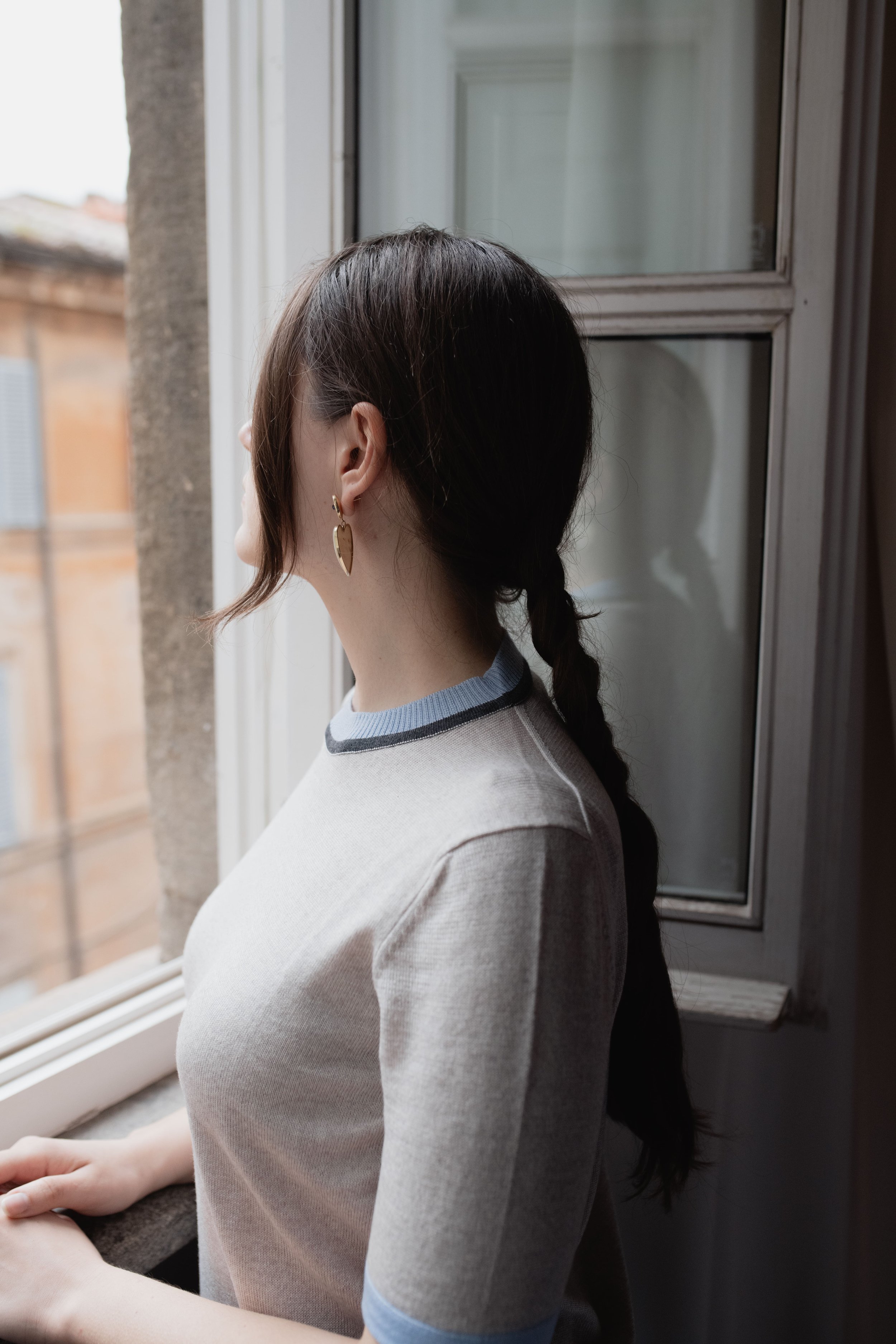Donna con capelli scuri raccolti in treccia guarda fuori dalla finestra in una stanza luminosa.