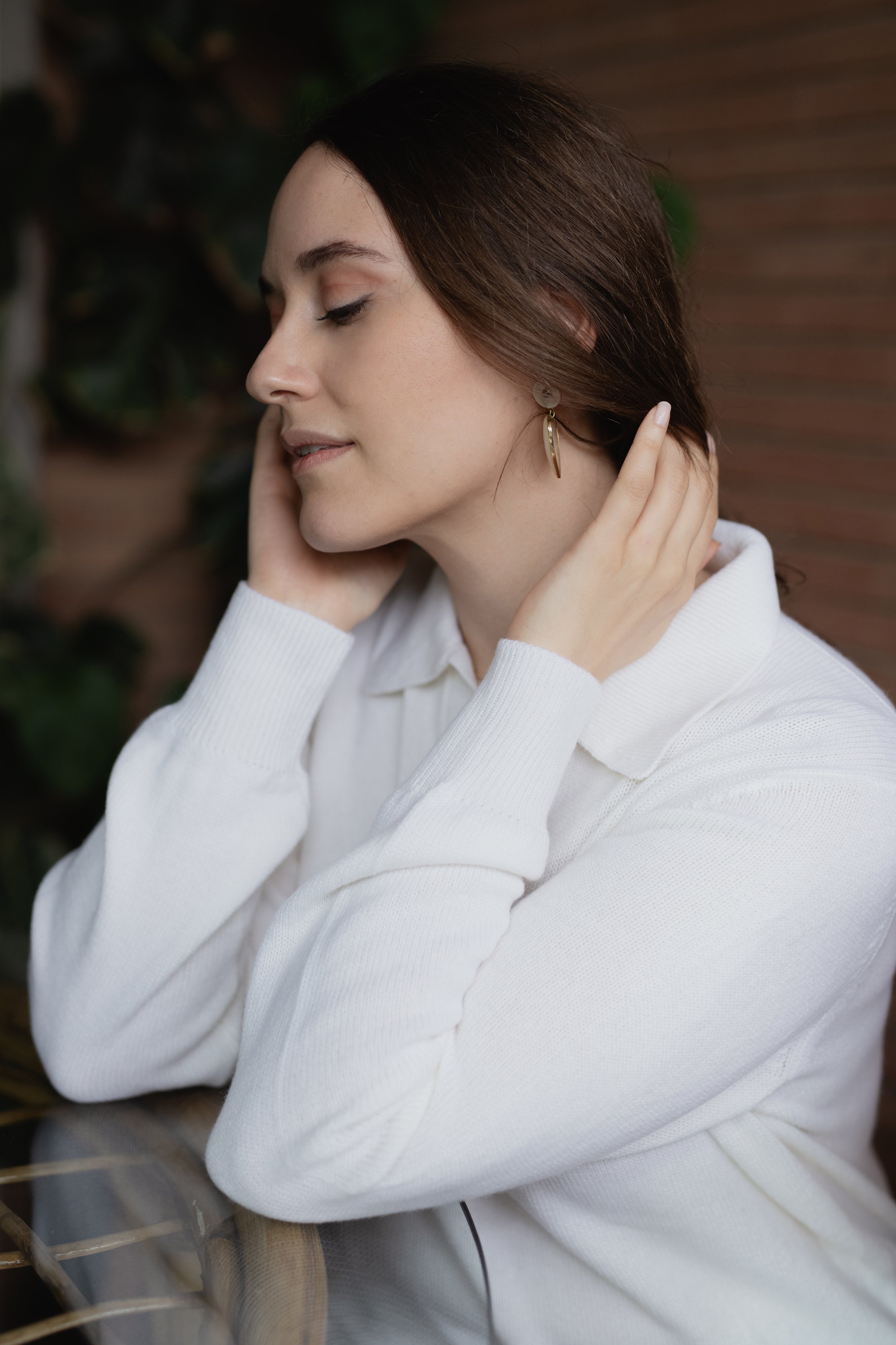 Donna con maglione bianco e orecchini dorati, rilassata con occhi chiusi, sfondo sfocato di foglie e mattoni.