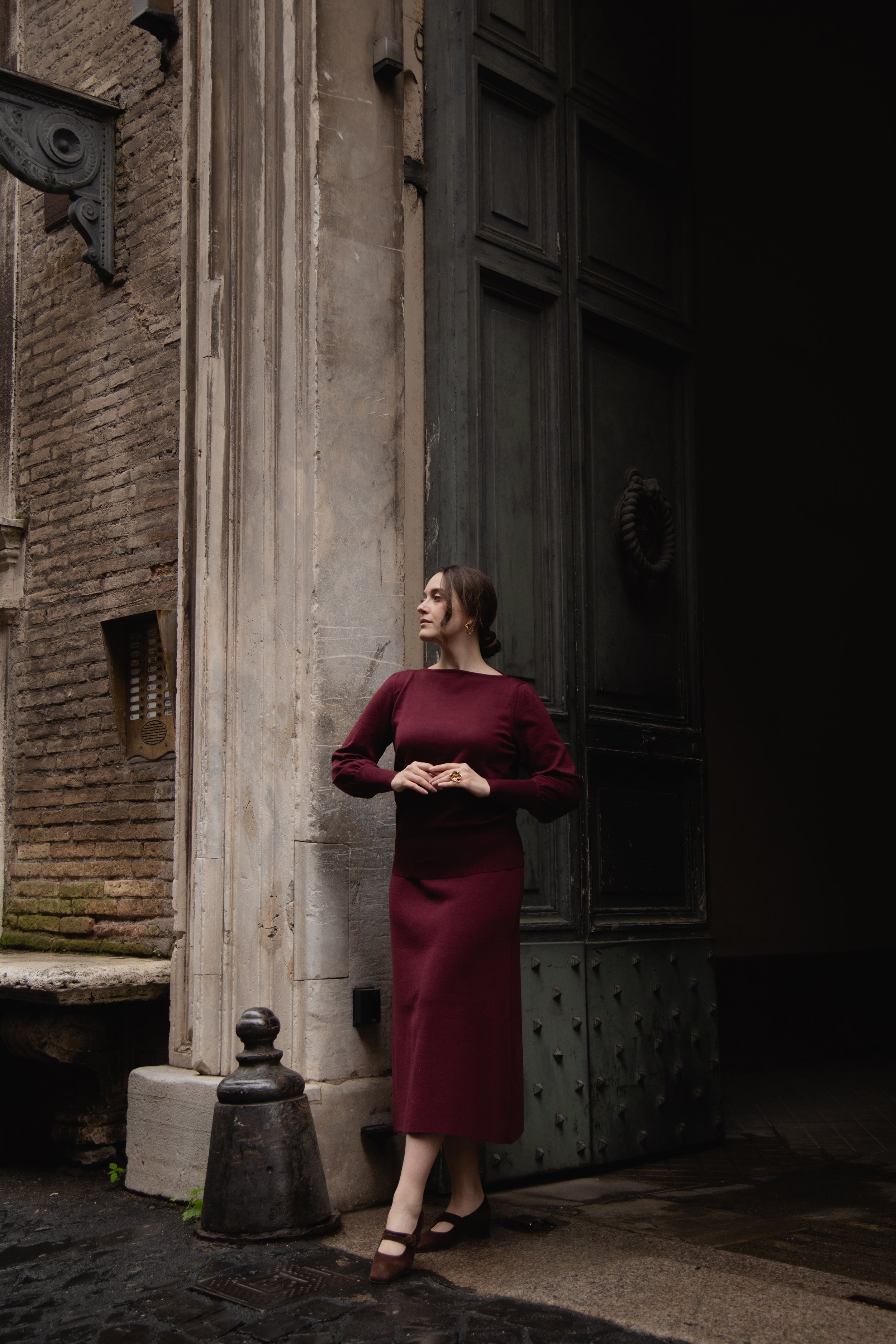 Donna in abito bordeaux in piedi accanto a una grande porta antica, sfondo architettura storica.