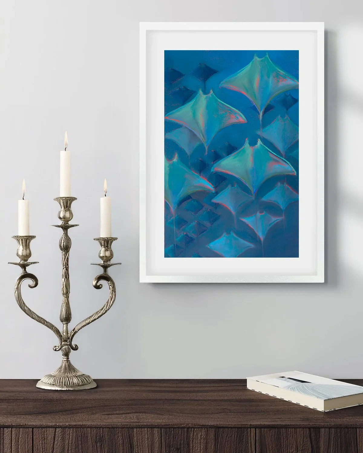 A framed abstract painting of blue and teal shapes on a white wall, a silver candelabra with three lit candles on a dark wooden table, and a closed white book.