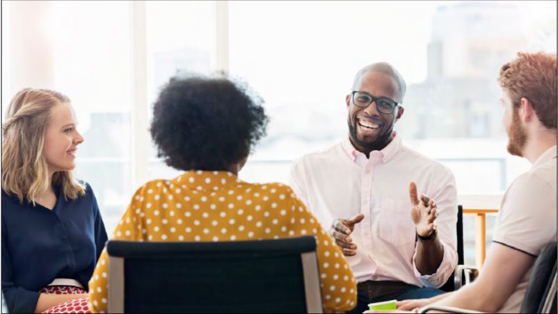 A group of people of mixed ages, genders and ethnicities sit round a meeting table having a discussion. The image represents a cognitively diverse group of people and the links between resilient leadership,  psychological safety and wellbeing