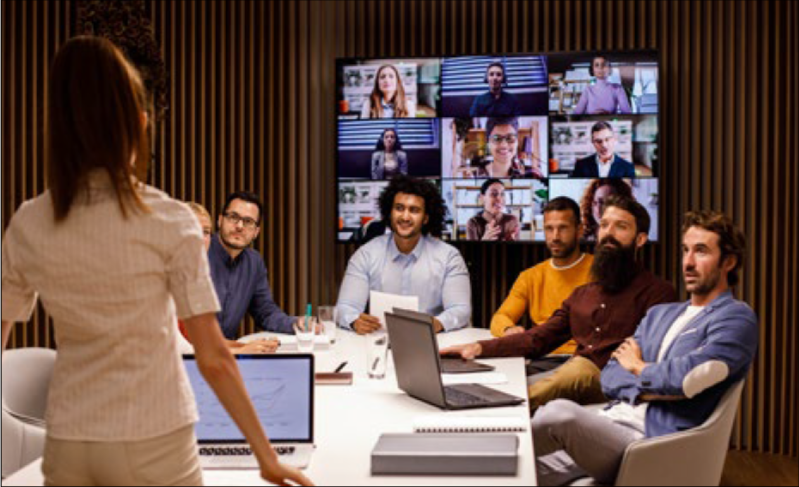 A woman stands presenting to a group of people some are dialing in via teams the others are sitting around a table. The image represents the challenges of working Hybrid