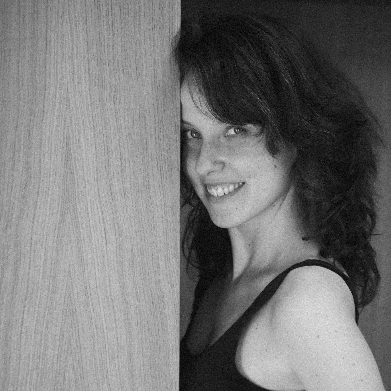 Black and white photo of a woman with dark wavy hair smiling, partially hidden behind a wooden panel.