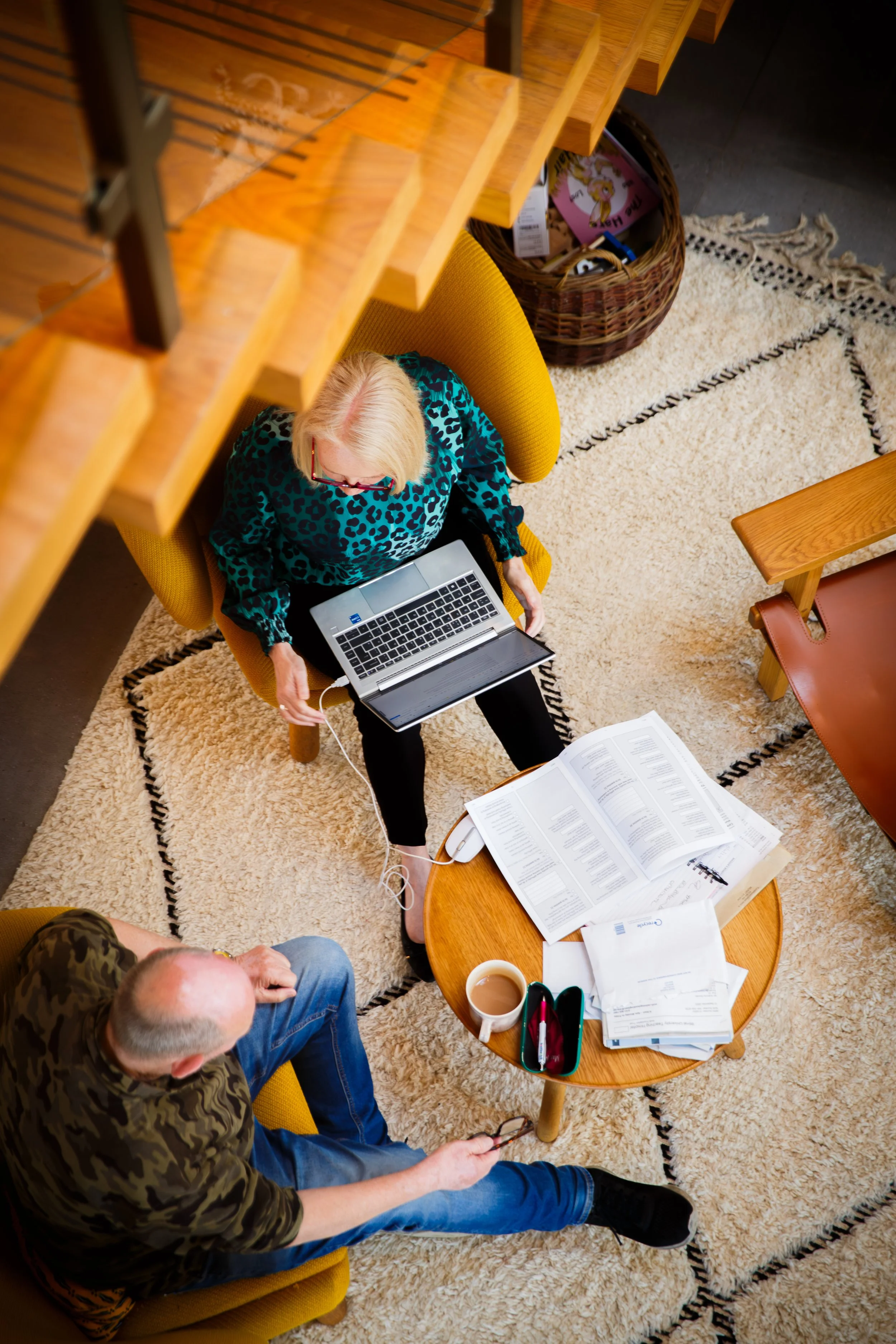 A woman sitting on a yellow chair using a laptop, a man sitting on a yellow chair holding glasses, a small wooden table with a cup of coffee, papers, and a pen. The scene is viewed from above, with a wooden staircase overhead.