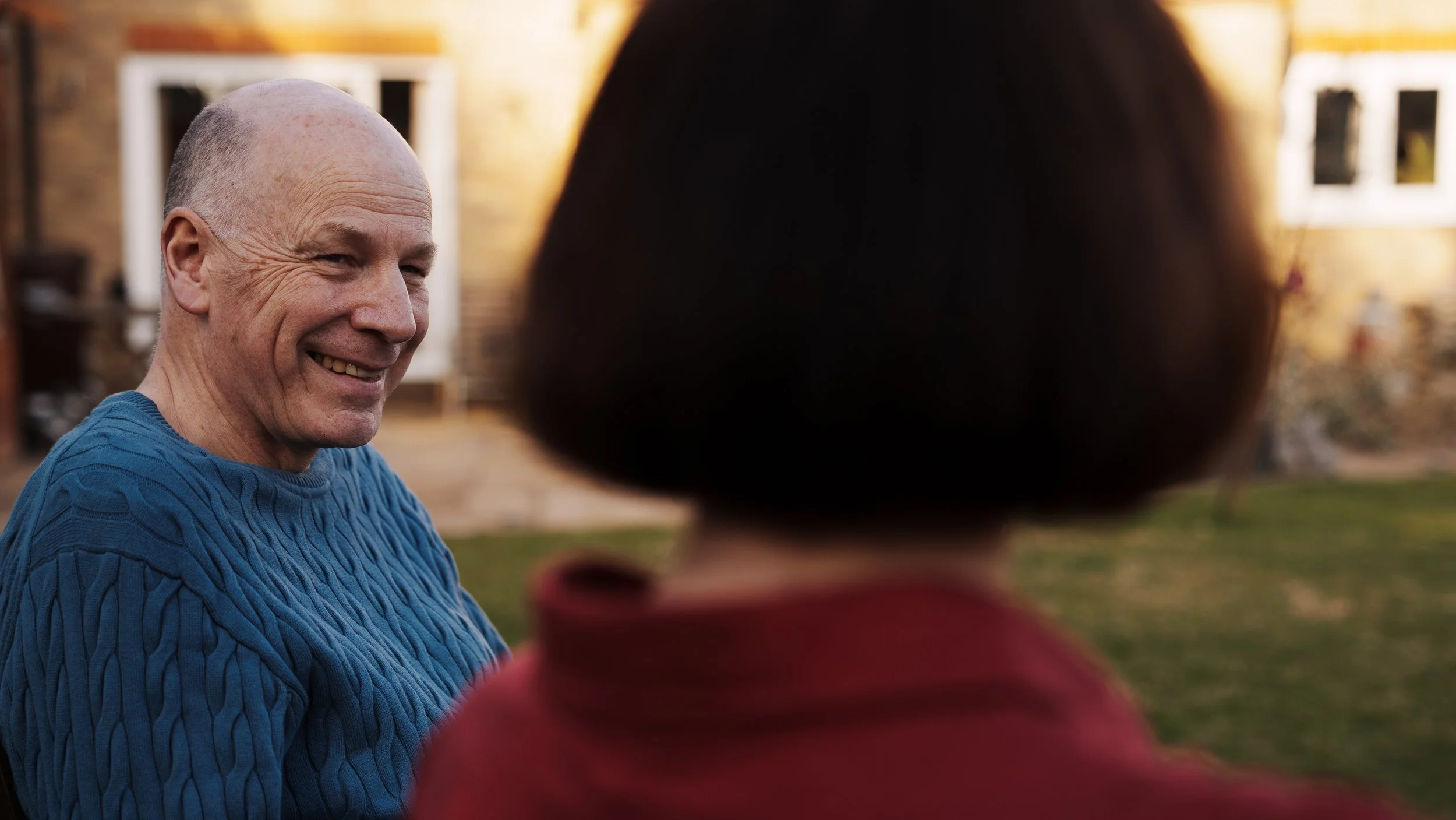 A smiling older man in a blue cable-knit sweater conversing outdoors with a woman whose back is to the camera, in their garden during sunset.