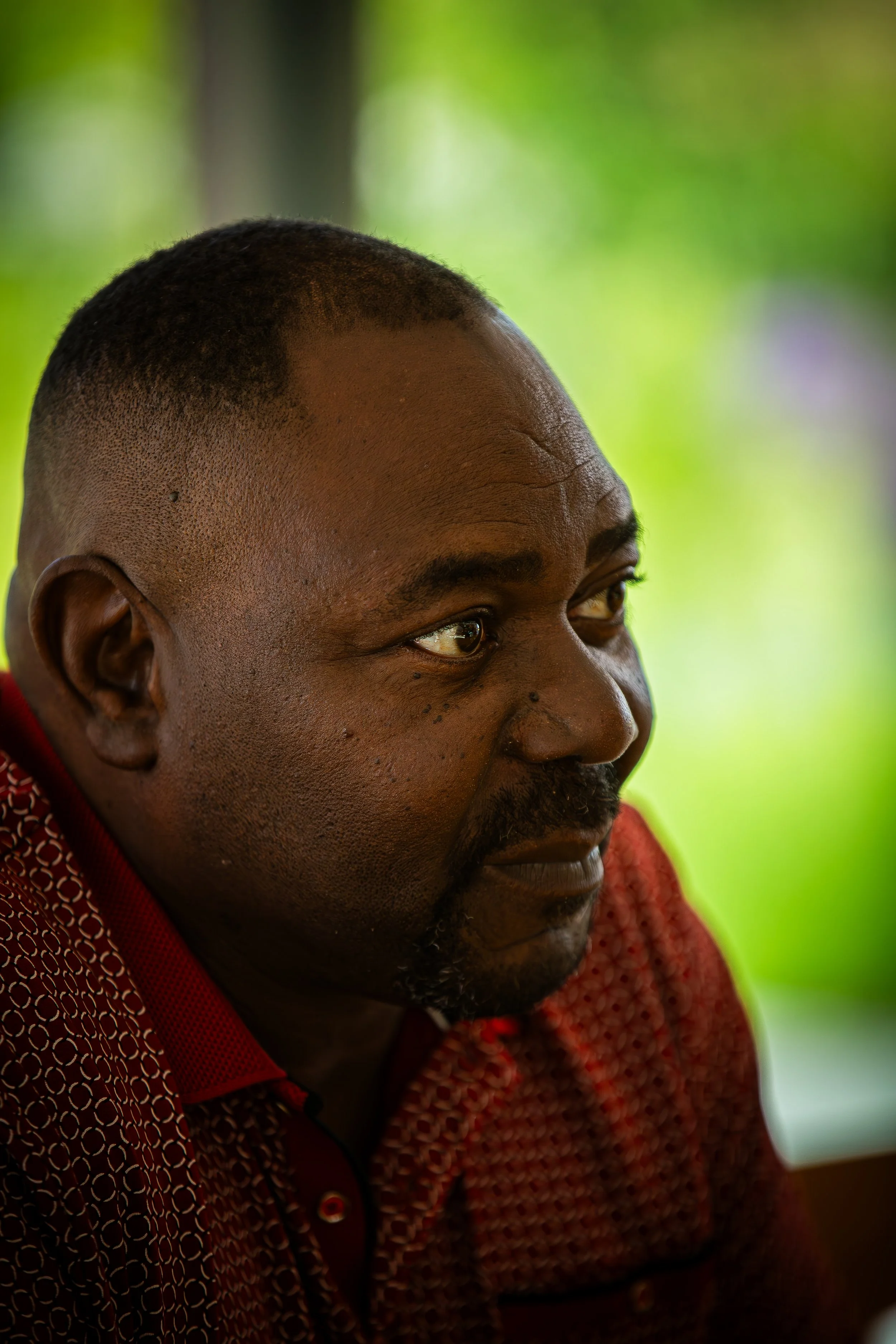 Close-up of a man with short hair and goatee wearing a red patterned shirt, looking to his left with a slight smile.