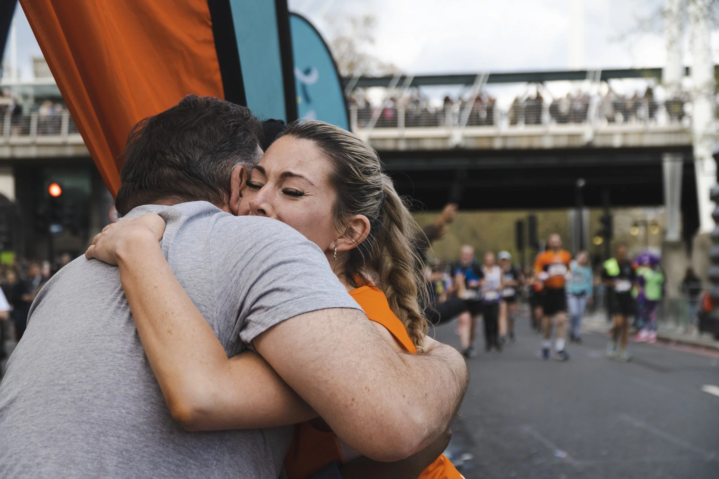 Two people hugging at a race event, with participants in the background during London Marathon.