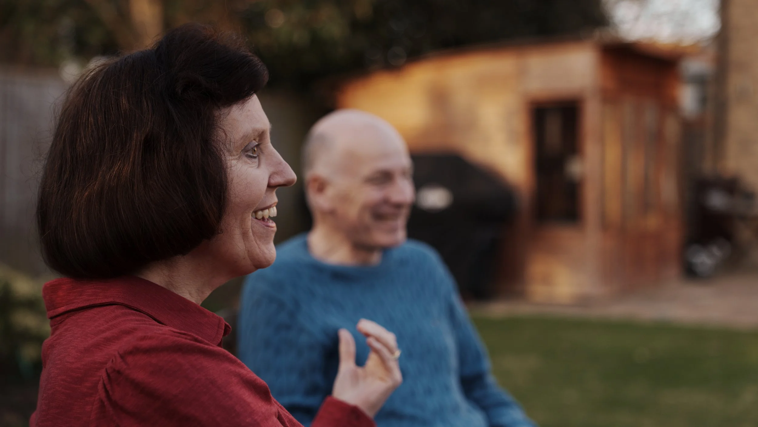 A woman with dark hair smiling in profile, wearing a red shirt, outdoors with a man with a shaved head in a blue sweater, smiling in the background.