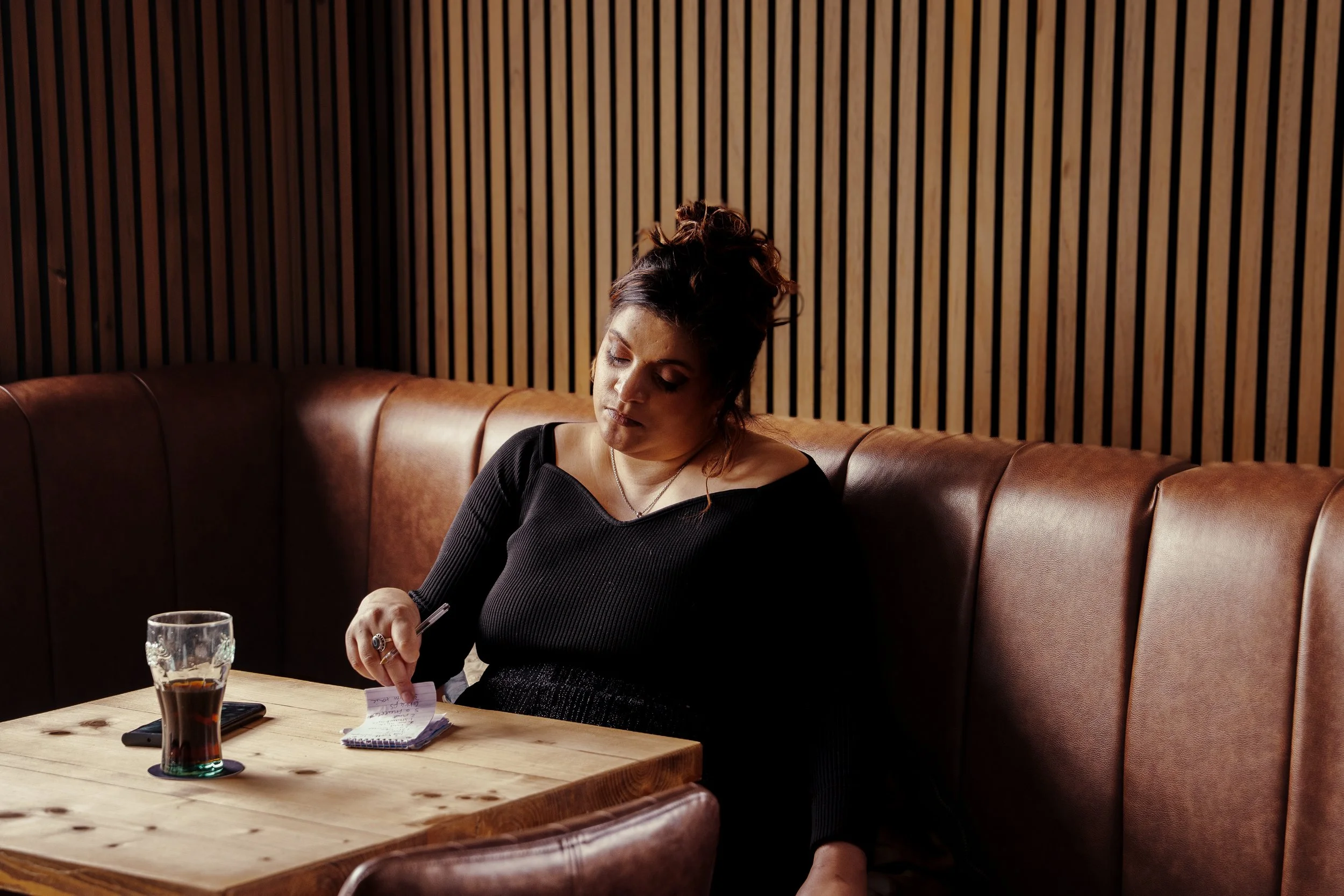 A woman with dark hair in a bun, wearing a black top, sitting on a brown leather bench, writing in a notepad on a wooden table, with a drink and a smartphone nearby, against a wooden slatted wall.