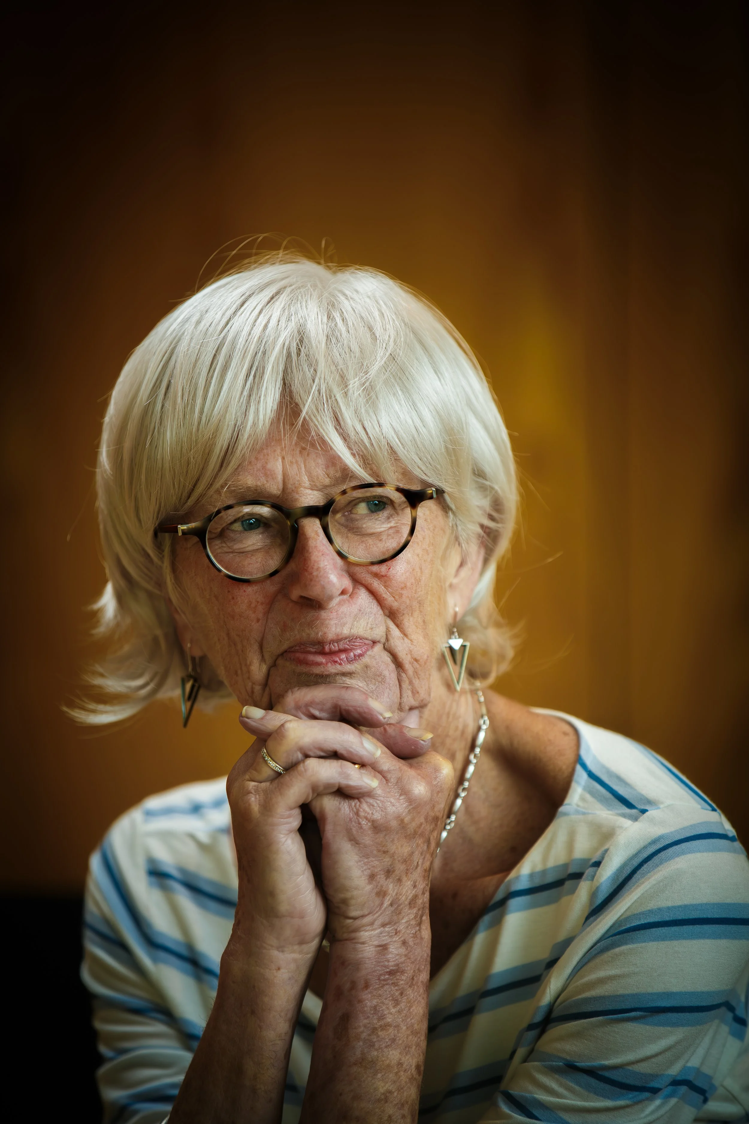 An older woman with white hair, wearing glasses, and a striped shirt, appears thoughtful with her hands clasped under her chin.