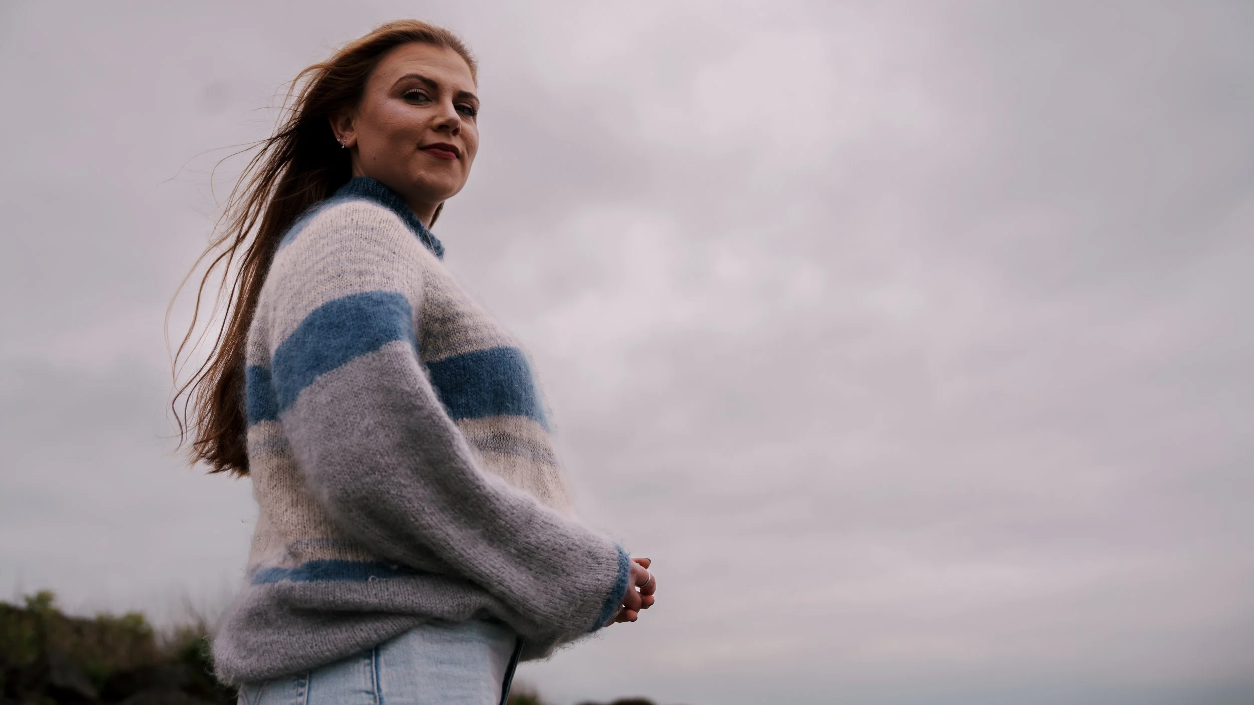 A woman with long red hair wearing a striped sweater standing outdoors under a cloudy sky.