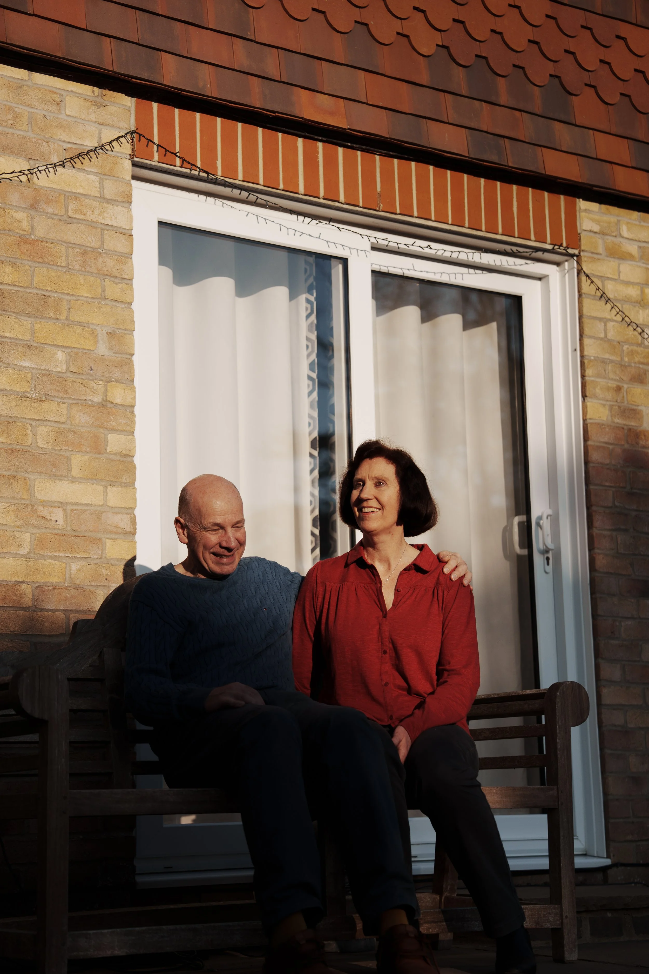 An older man and woman sitting together on a wooden bench outside a brick house, smiling and enjoying each other's company during sunlight.