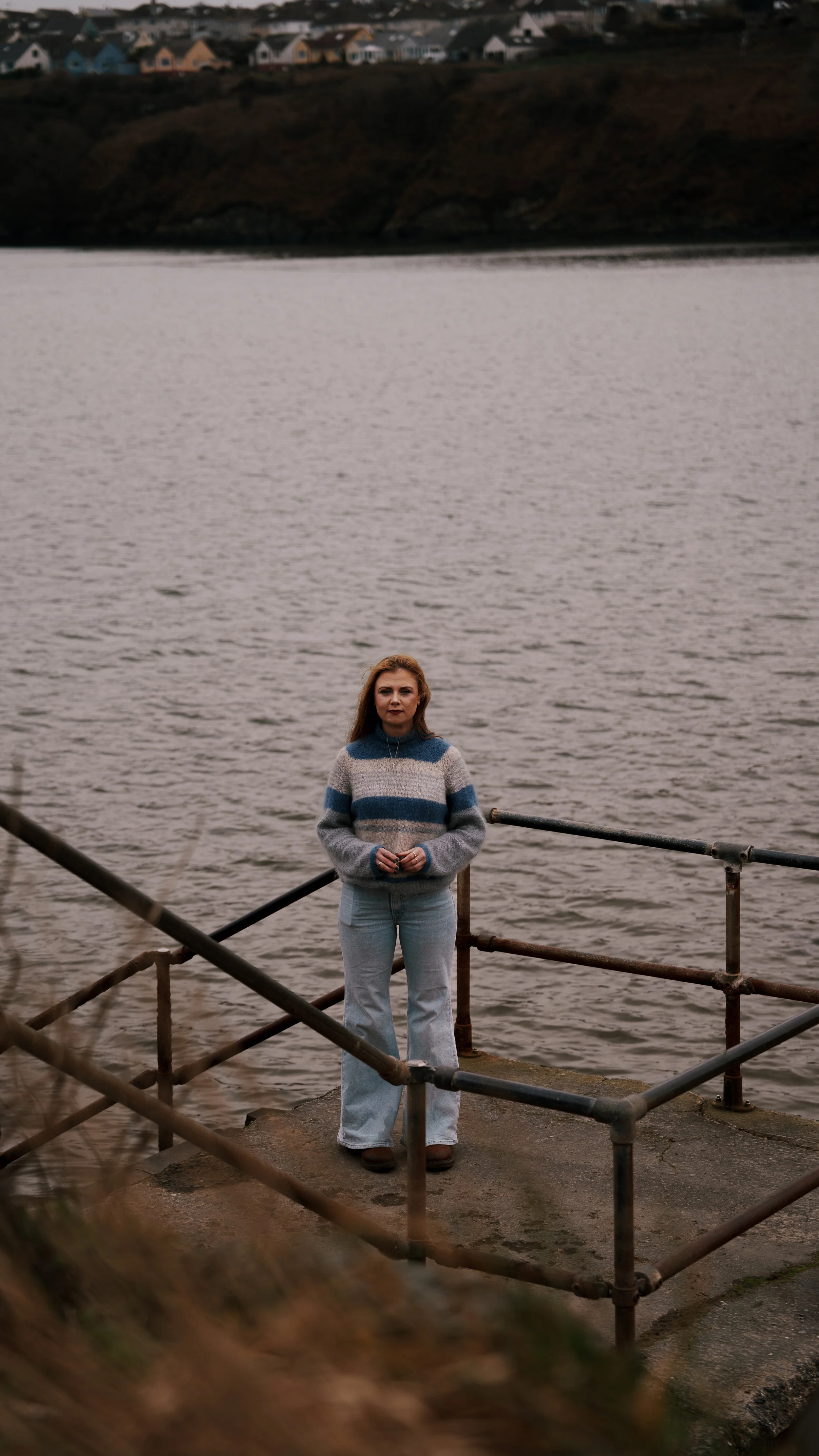 A woman standing on a concrete platform by a body of water, with houses on a hill in the background.