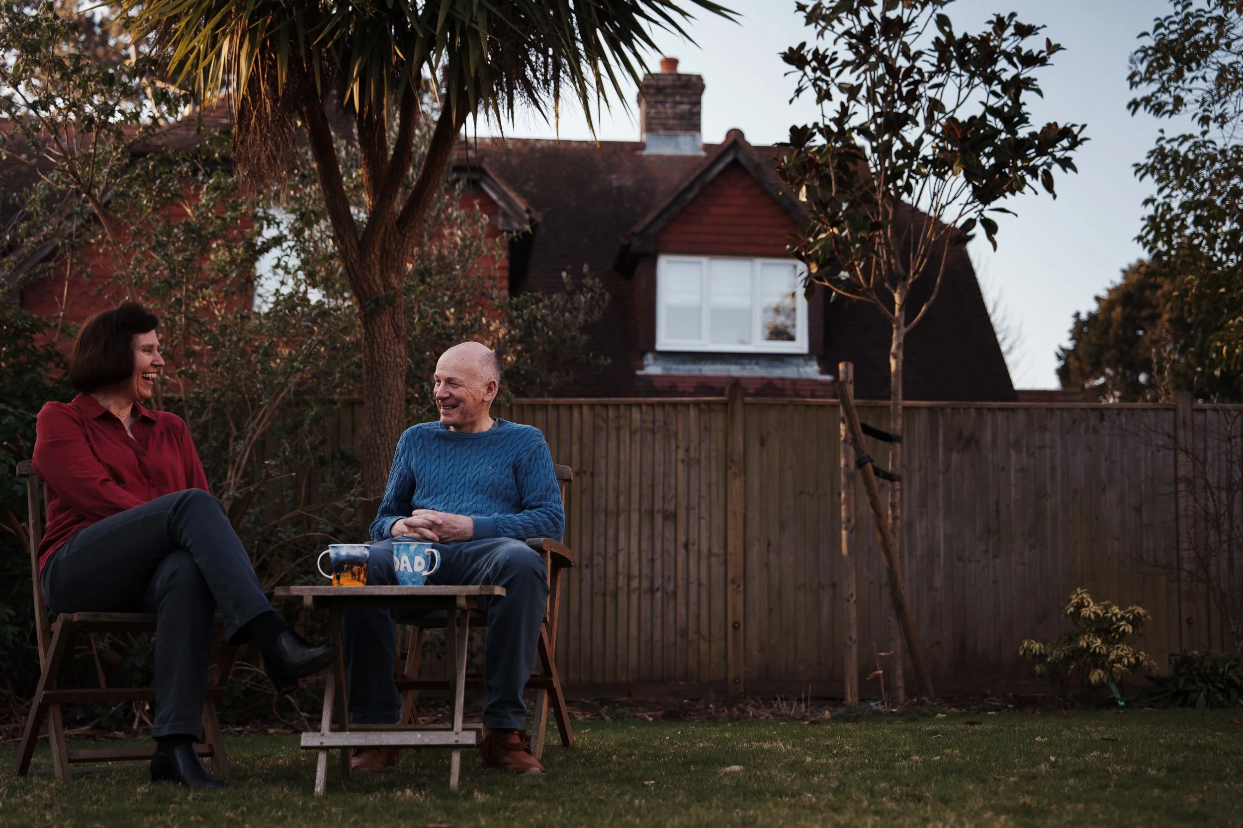 A woman and a man sitting on lawn chairs in a backyard, smiling and talking, with a house, trees, and a wooden fence in the background during dusk.