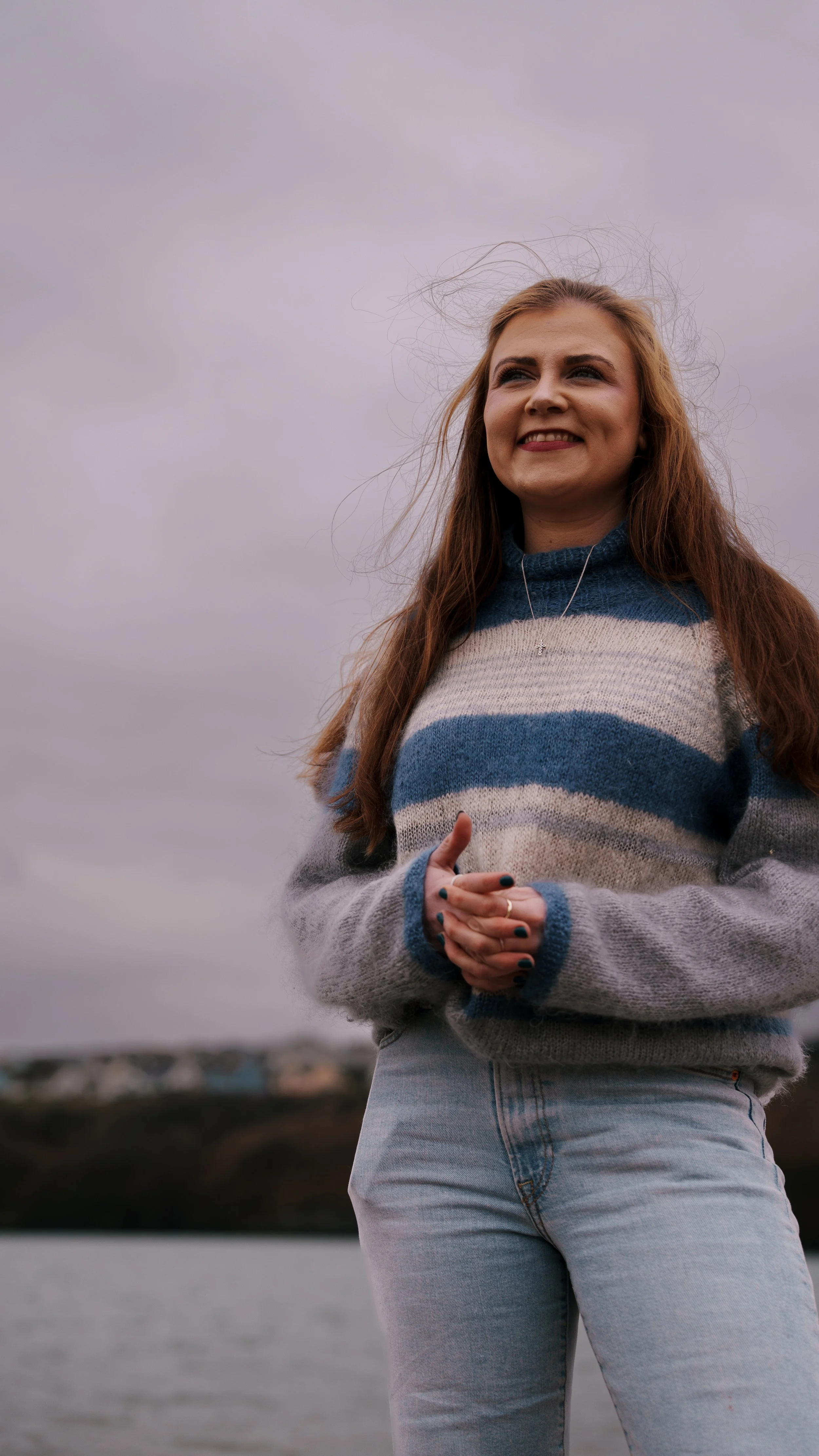 A young woman with long red hair wearing a striped sweater and light jeans stands outdoors near water, smiling under a cloudy sky.