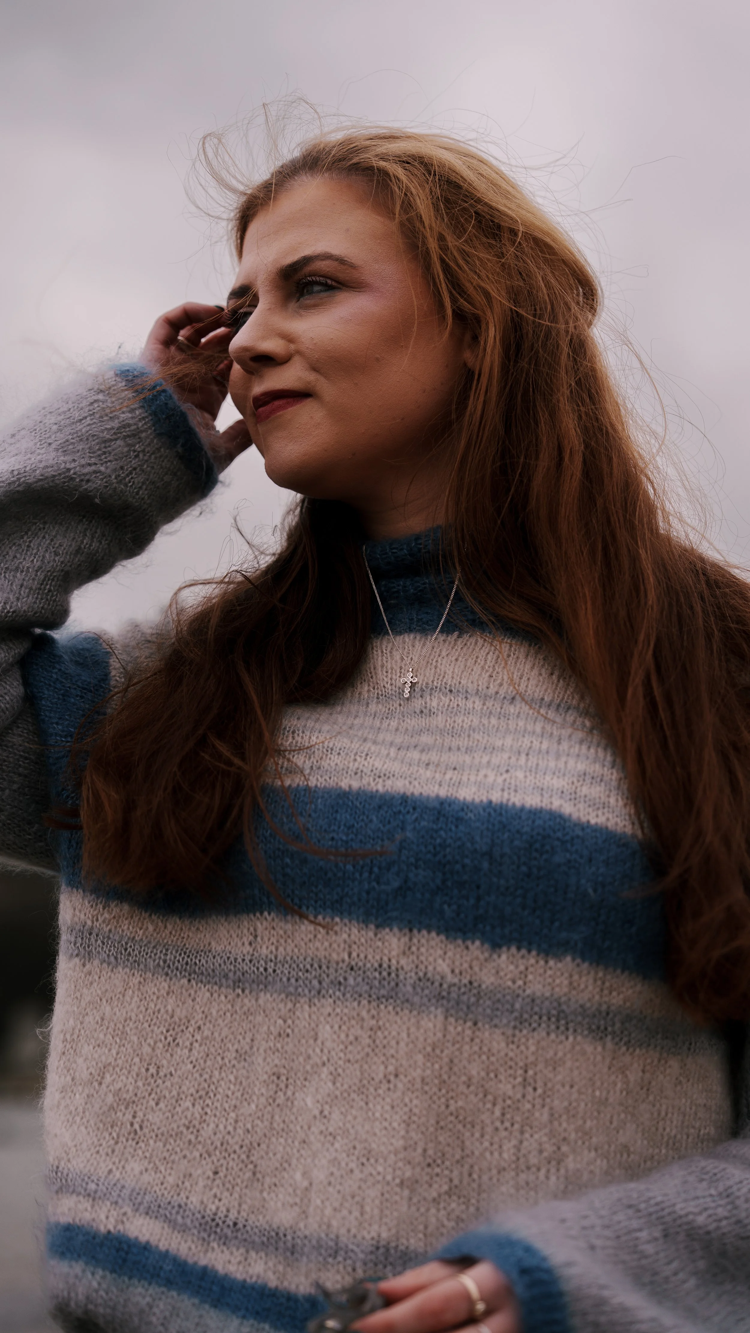 A woman with long red hair, wearing a striped sweater and a necklace, stands outdoors on a cloudy day, with her hand near her ear and a smile on her face.