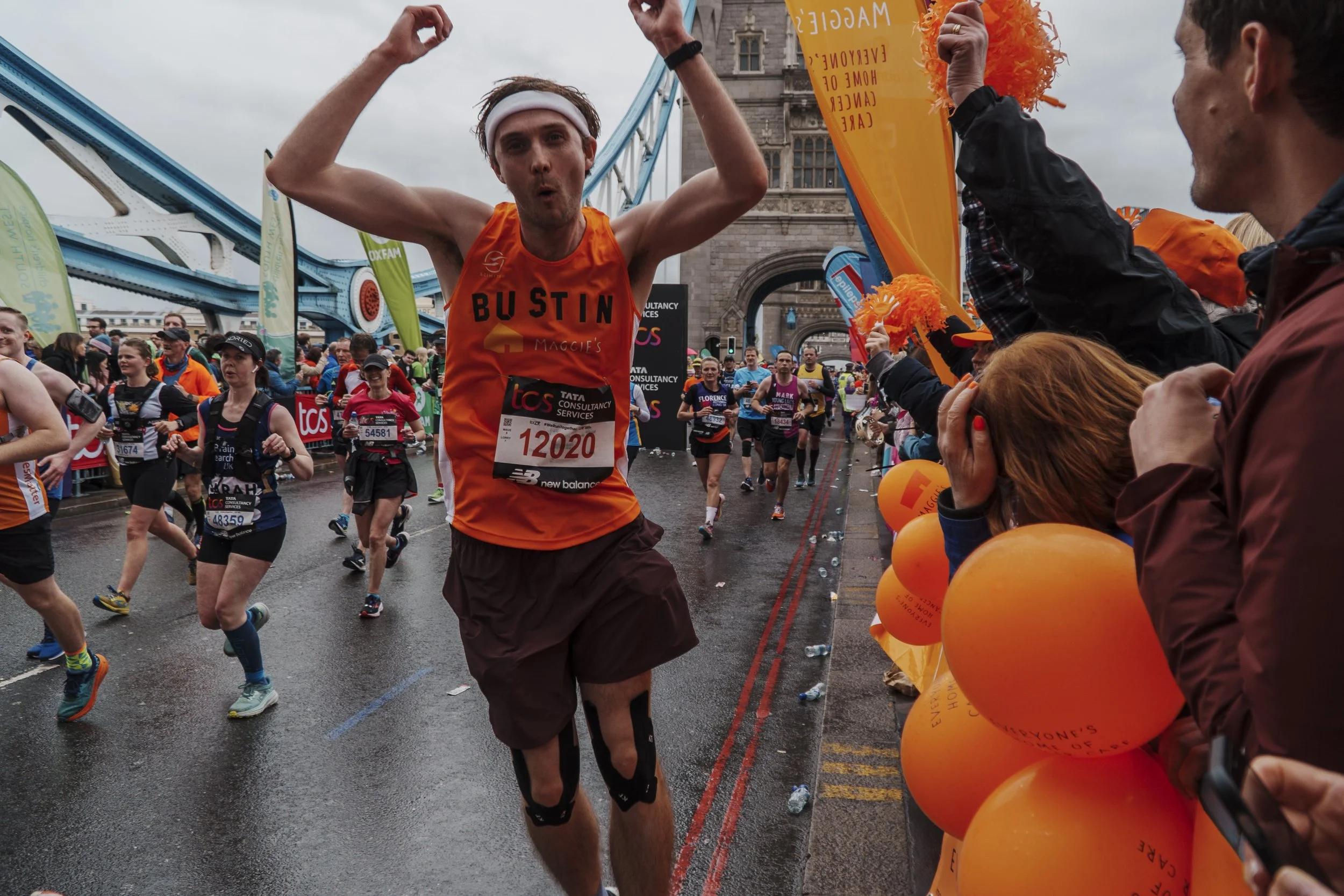 A male marathon runner wearing an orange tank top and brown shorts celebrating with the crowd, who are cheering and waving orange balloons along a rainy street with Tower Bridge in the background.