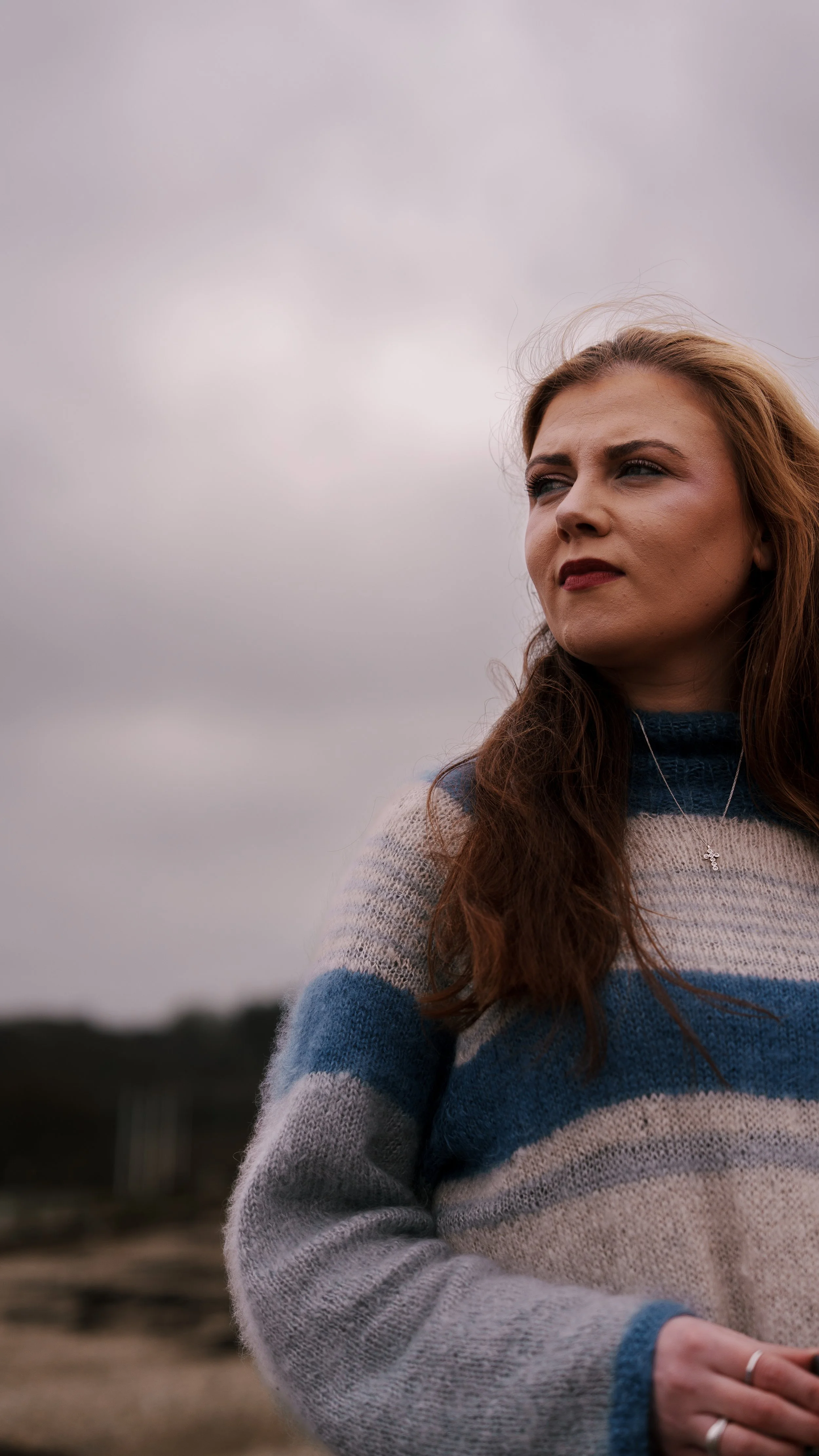 A woman with long red hair standing outdoors under a cloudy sky, wearing a striped sweater and a necklace.