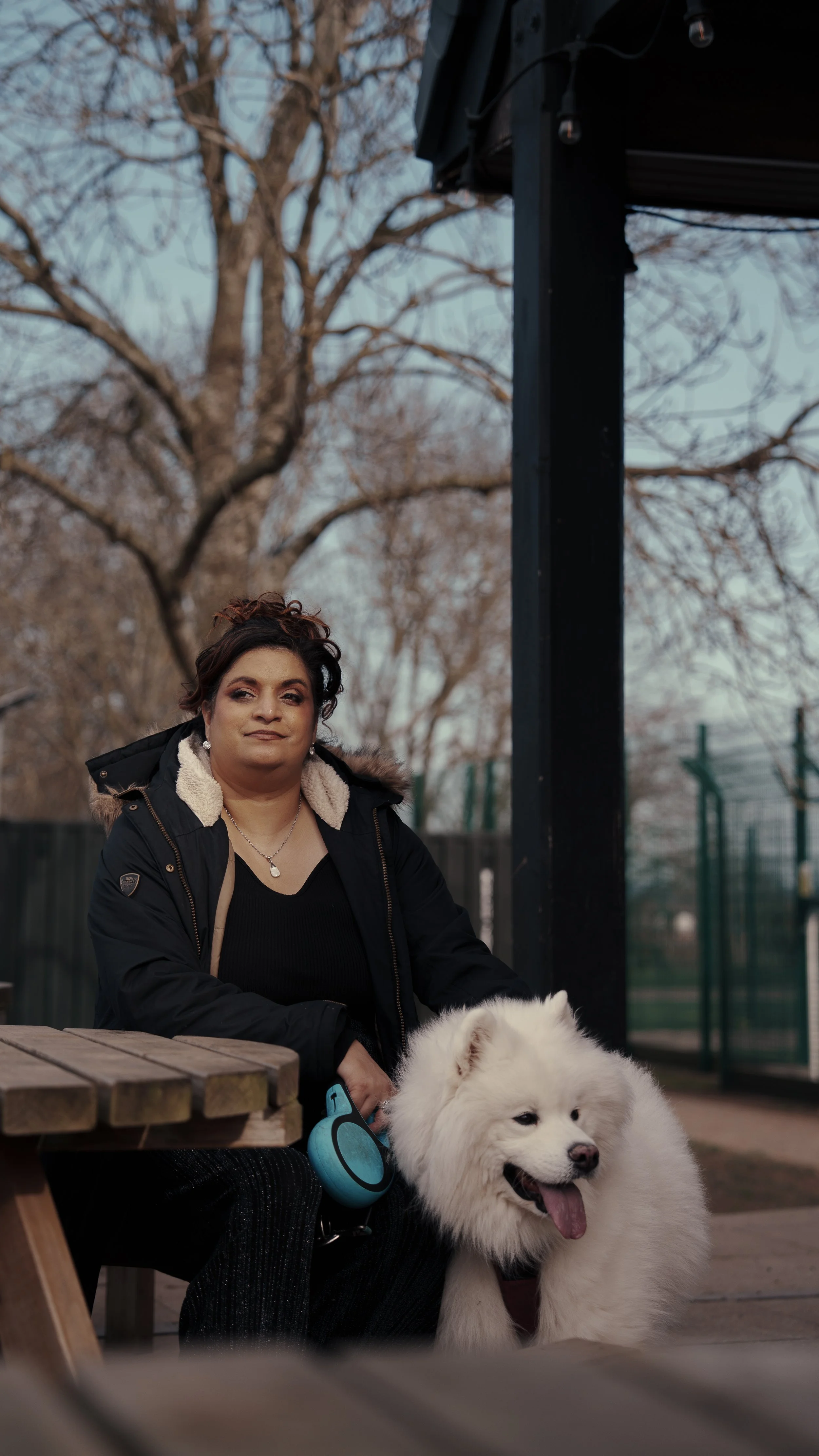 A woman sitting on a bench in a park with a large white fluffy dog at her side. The woman is wearing a black jacket with a fur-lined hood and has short curly hair. The background includes leafless trees and a fence.