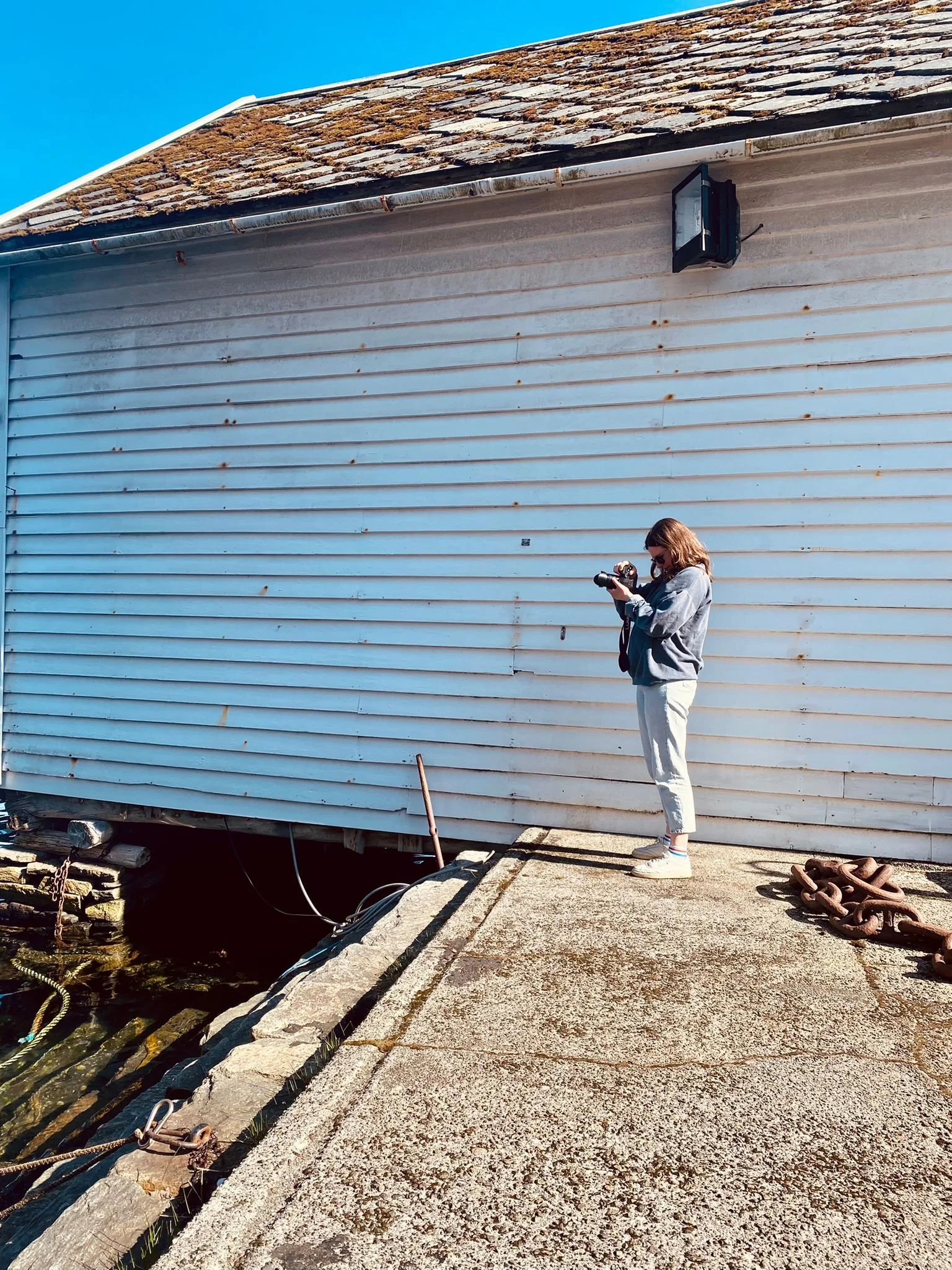 A woman standing on a concrete dock taking photos with a camera, next to a white building with horizontal siding and a stone roof, near a water body.