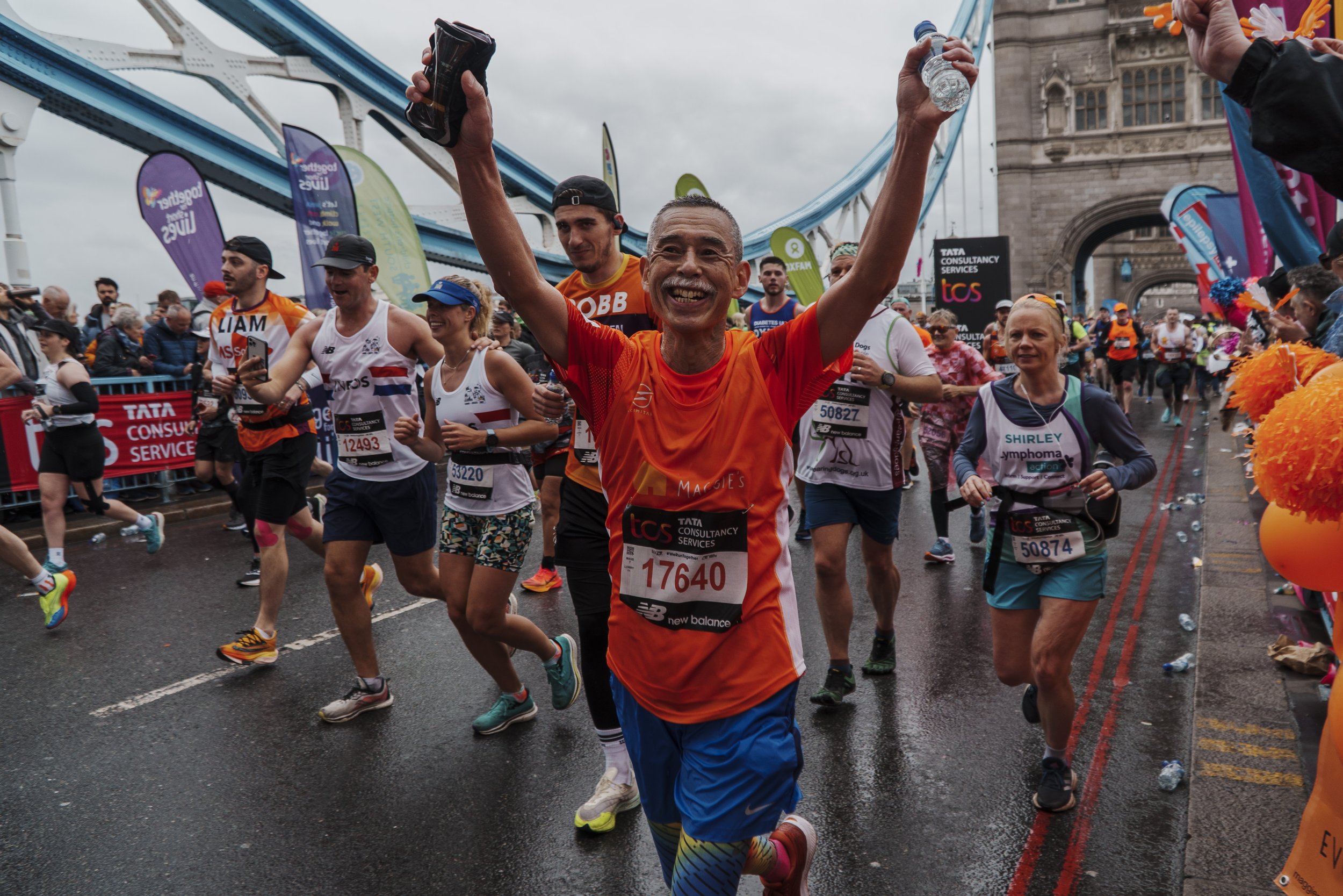 A group of marathon runners running over Tower Bridge with the central runner, holding a water bottle, smiling and celebrating. 
