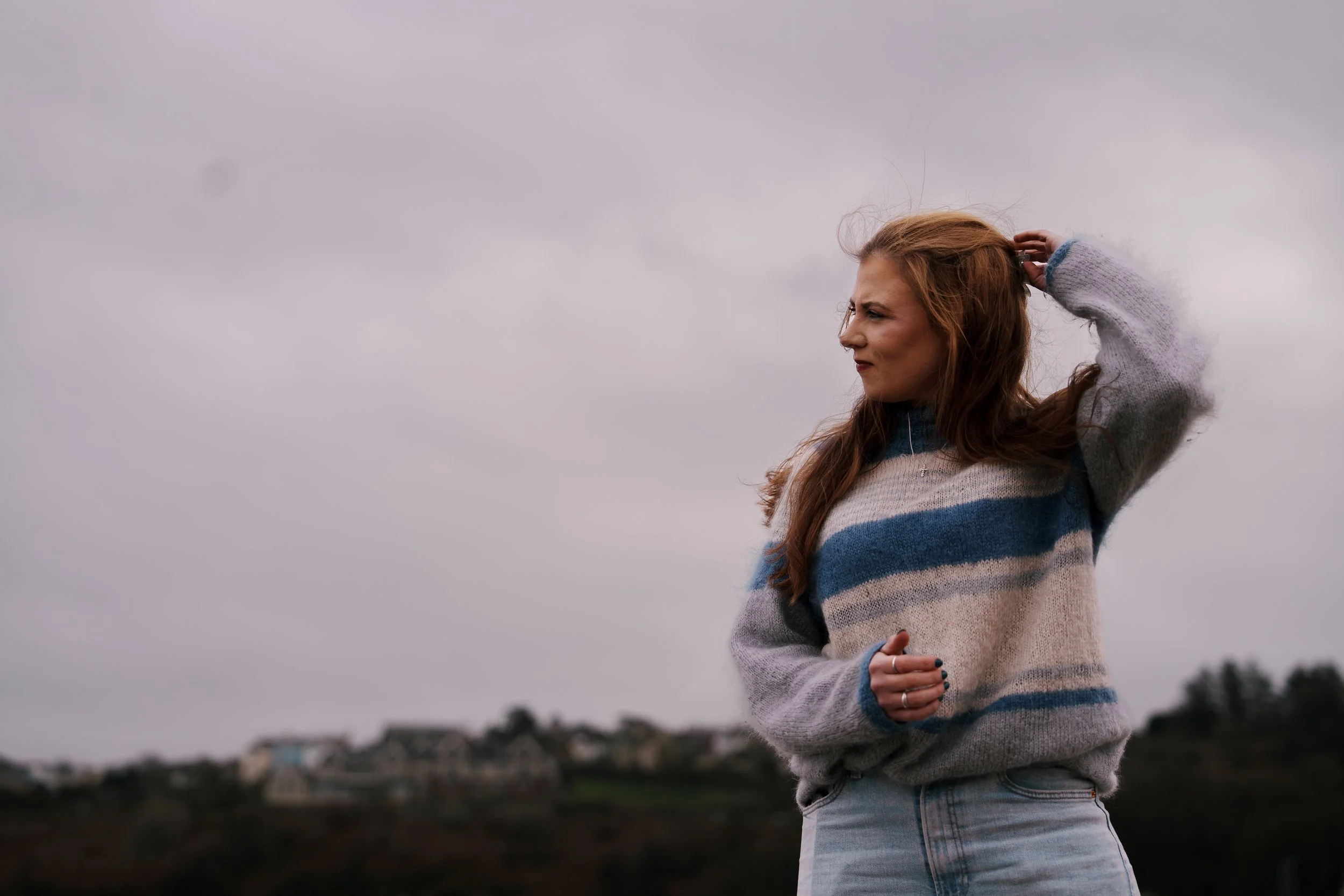 A young woman with long red hair standing outdoors under a cloudy sky. She is wearing a cozy striped sweater and light denim jeans. Her left hand is raised to her head, and she is looking to the side with a contemplative expression. In the background