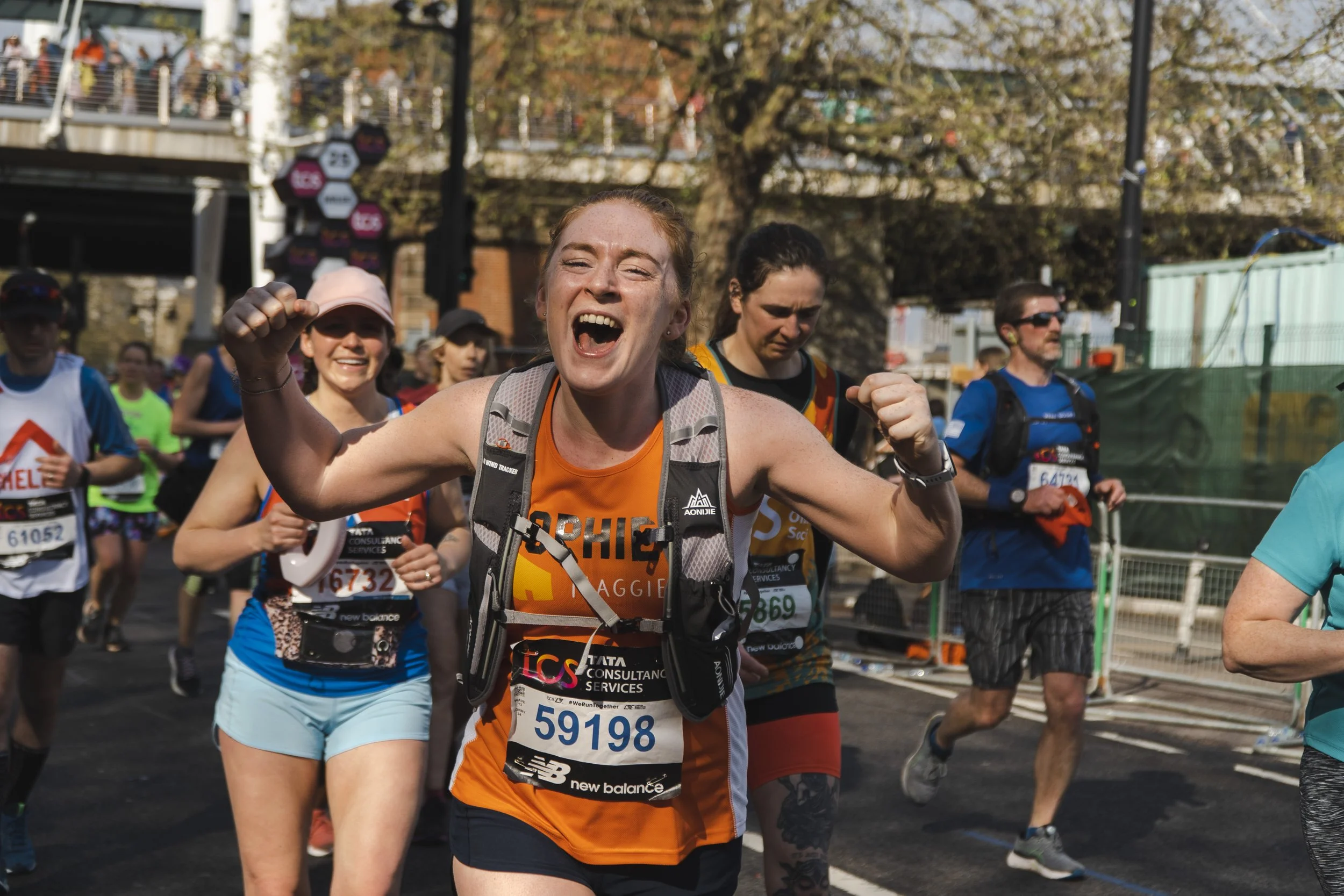 A group of marathon runners participating in a race, with a female runner in the foreground showing a victorious pose, smiling with clenched fists, wearing athletic gear and a race bib.