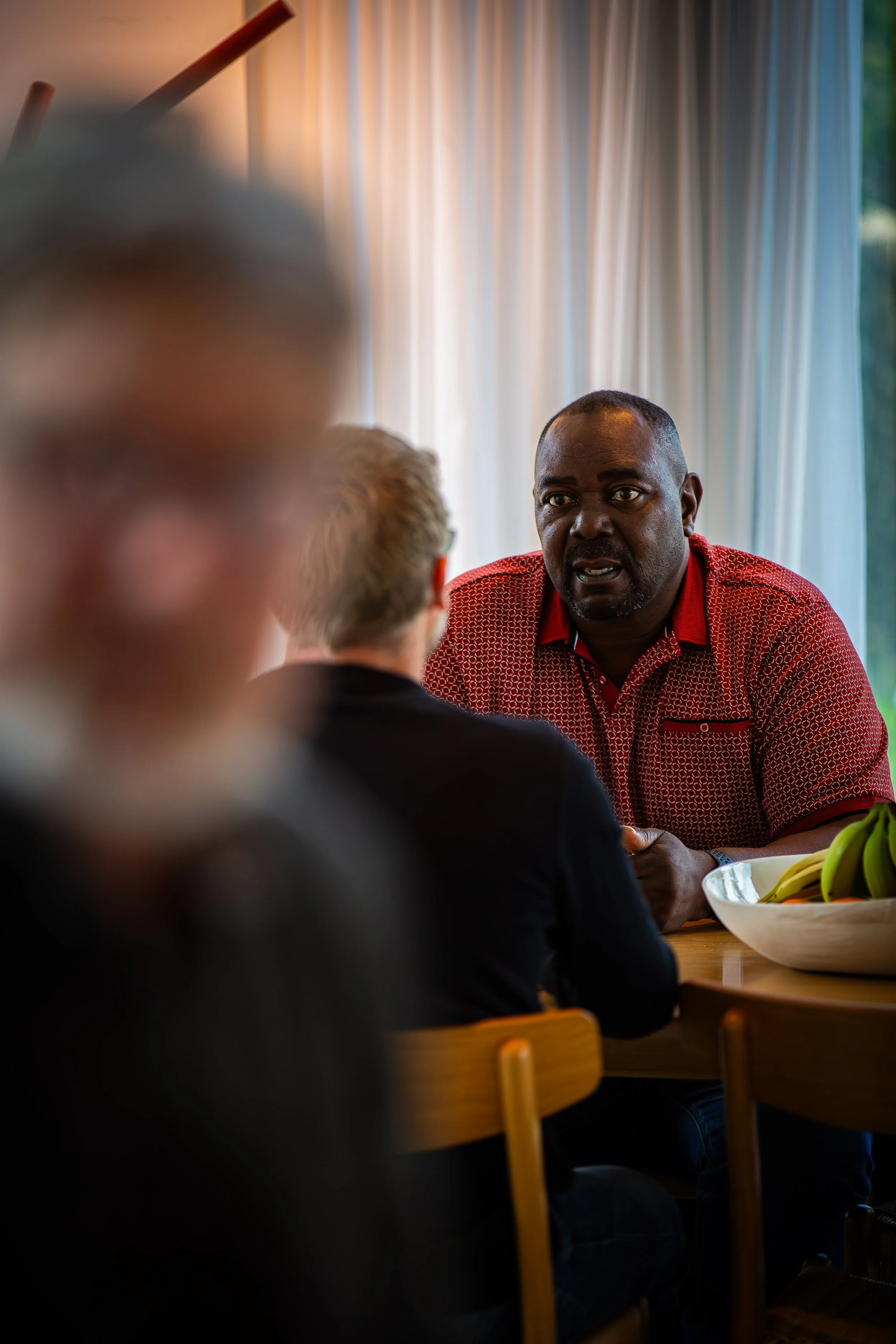 Man in red patterned shirt talking to a man in a black shirt with his back to the camera, with a blurred figure in the foreground.