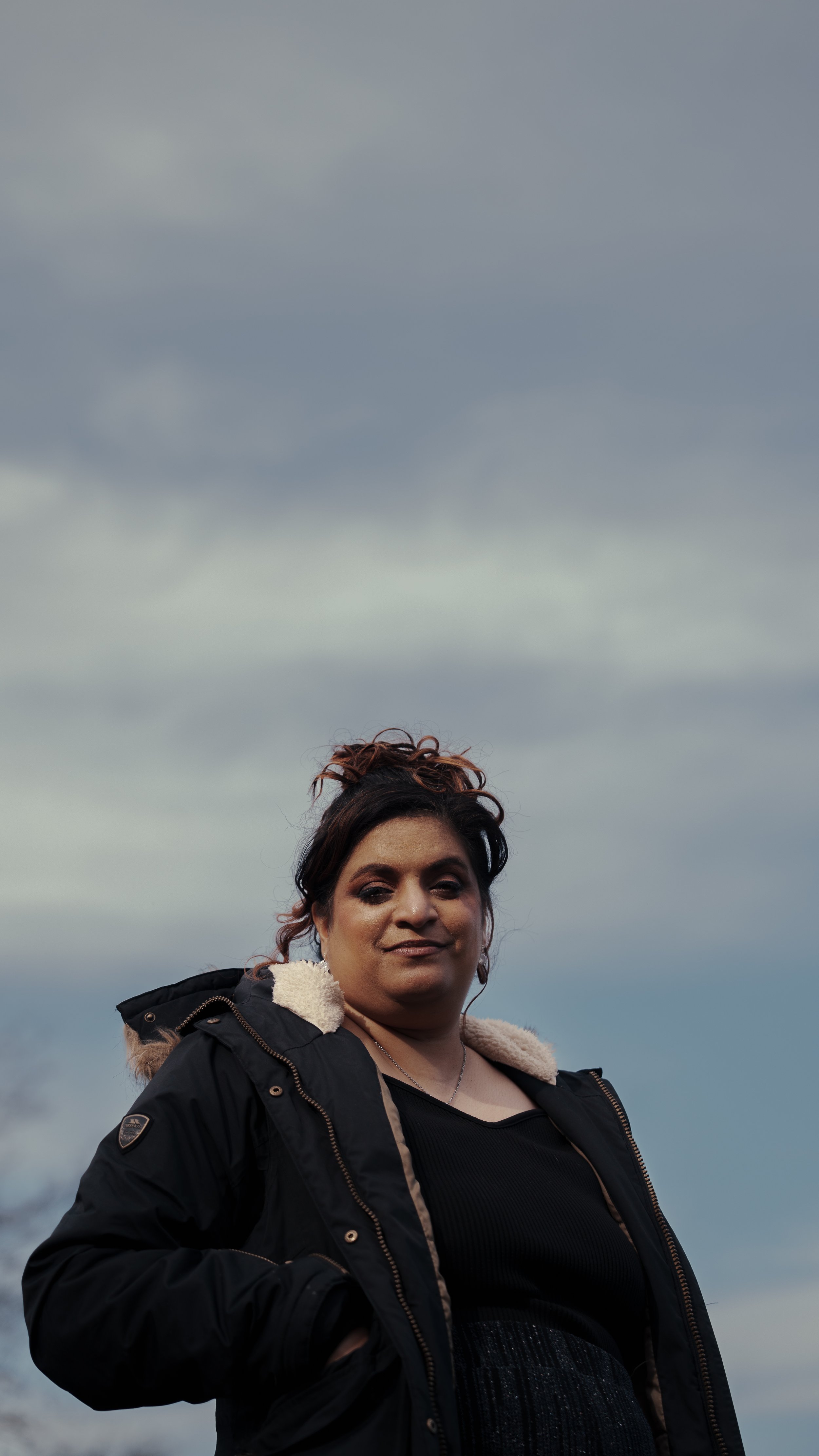 A woman with curly brown hair in an updo, wearing a black jacket, standing outdoors against a cloudy sky.