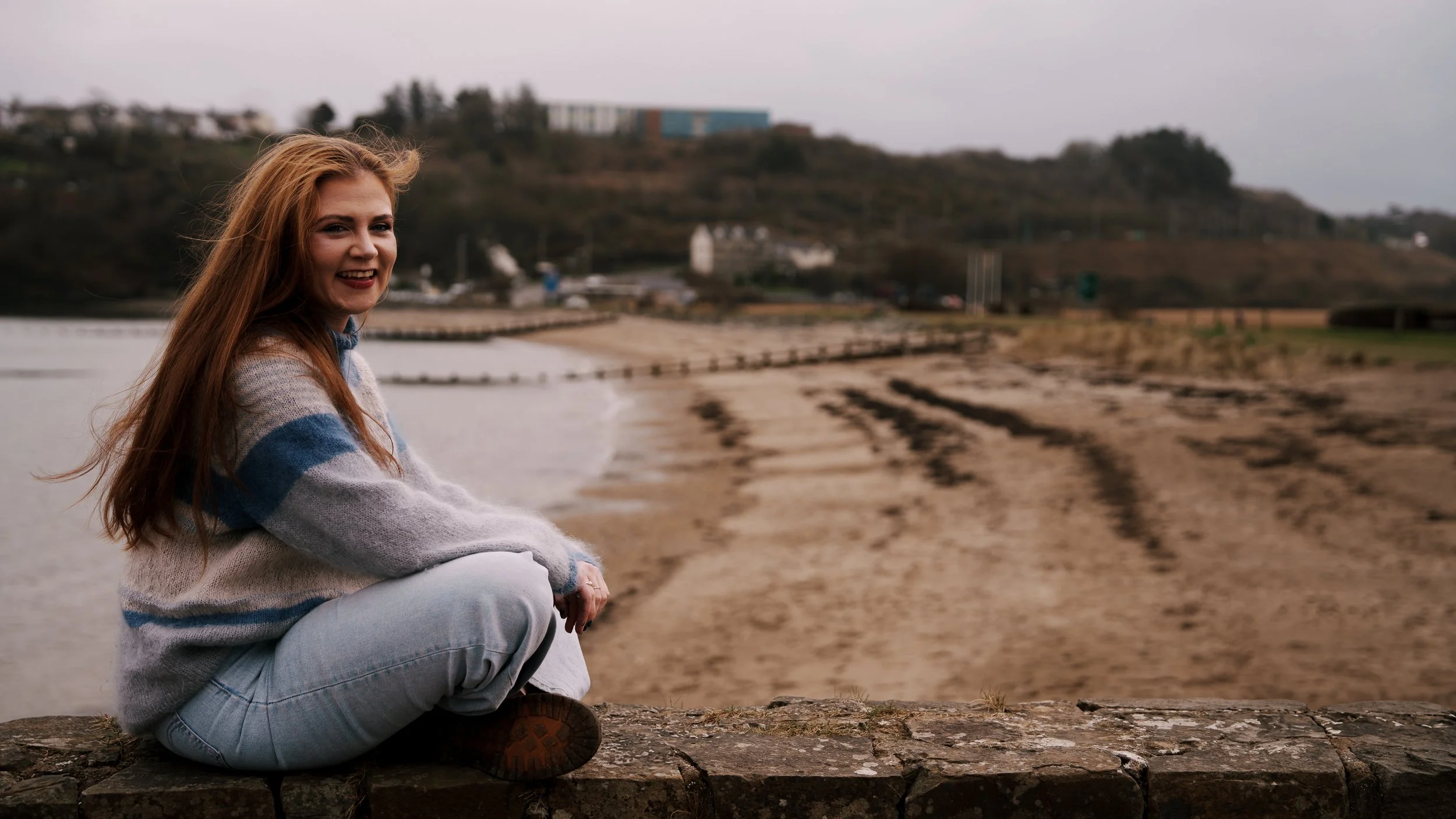 A young woman with long red hair, wearing a striped sweater and jeans, sitting on a stone wall by a beach, smiling at the camera during overcast weather.