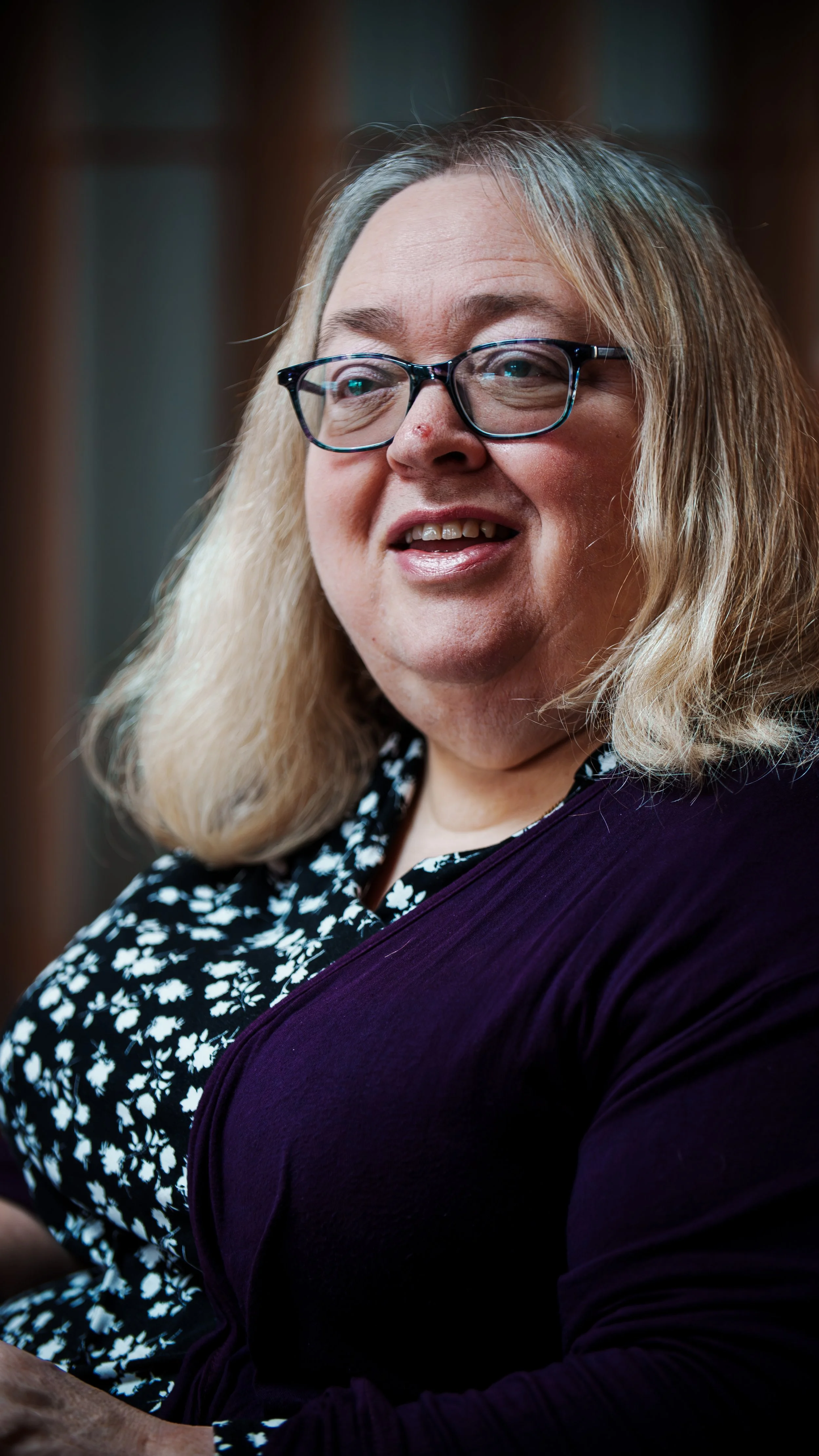 A woman with blonde hair, glasses, and a black floral blouse, smiling and looking to her left.