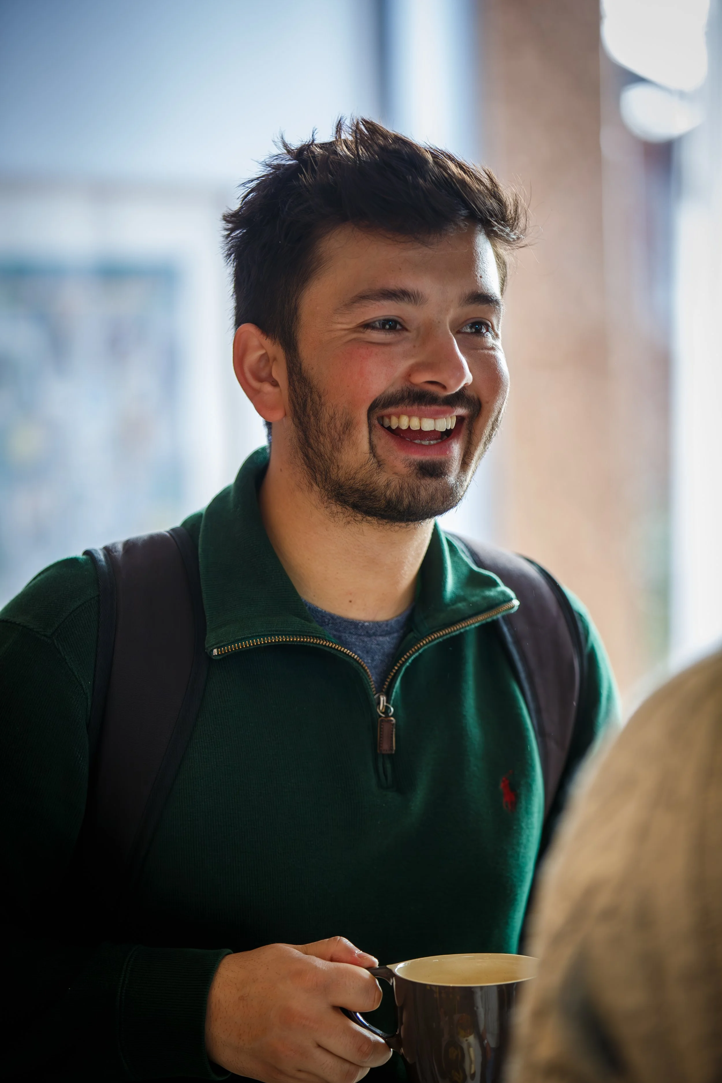A young man with dark hair and beard, smiling and holding a coffee cup, wearing a green pullover.